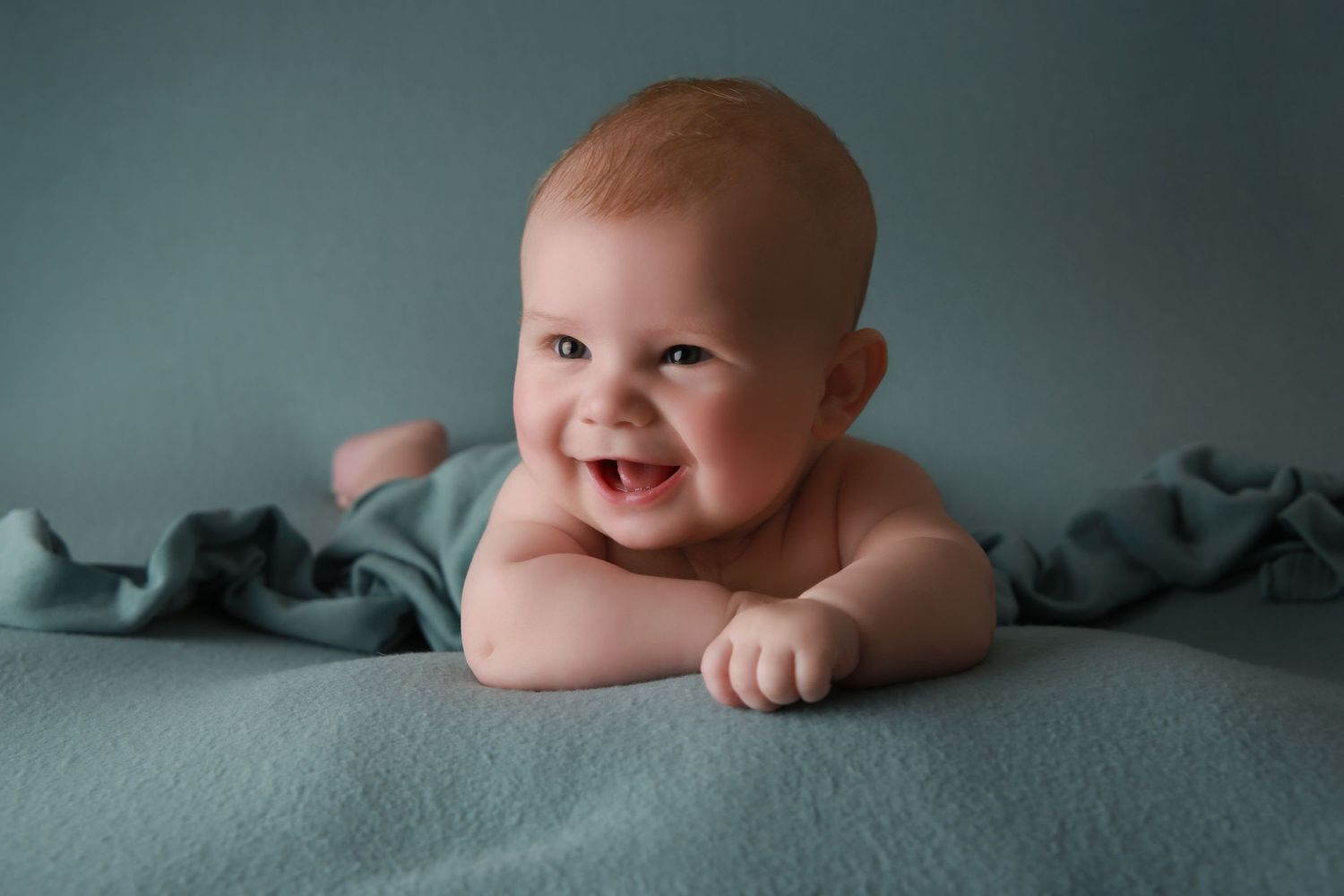 Smiling baby with blue eyes lying on teal blanket, cheerful infant portrait photography.