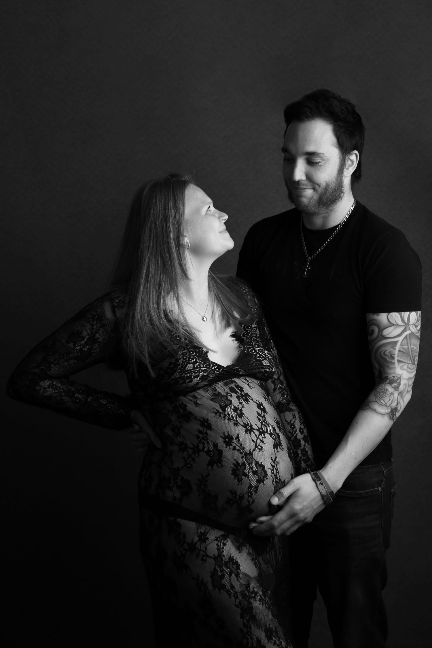 Black and white maternity portrait of tattooed man and pregnant woman in lace dress.