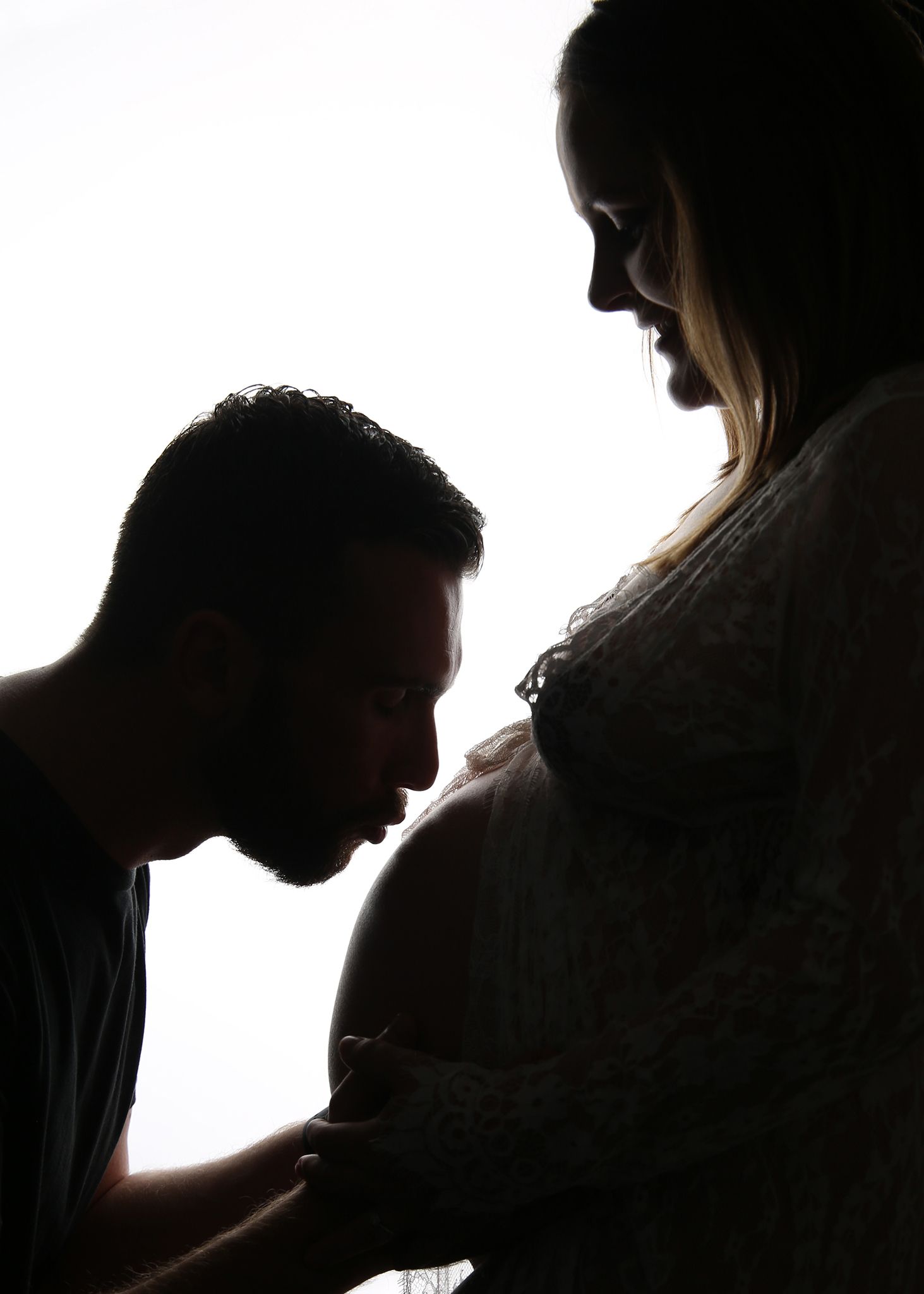 Silhouette of a man kissing a pregnant woman's belly against a bright white background.
