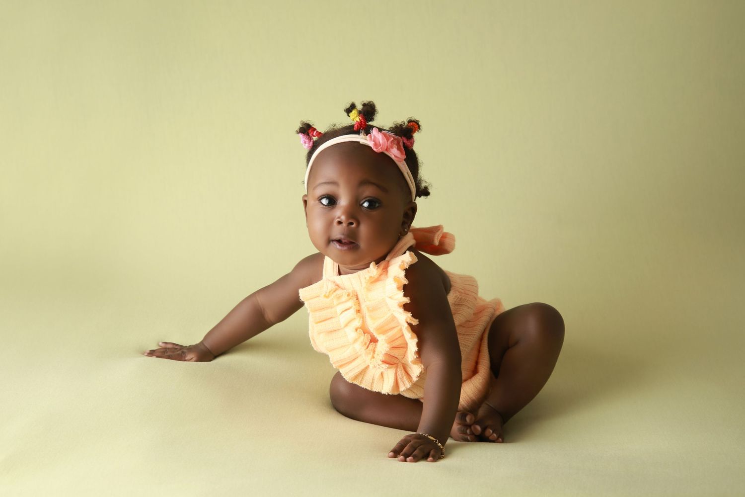 A cute baby girl in an orange ruffle romper sits on a green background with colorful pom-pom hair accessories.