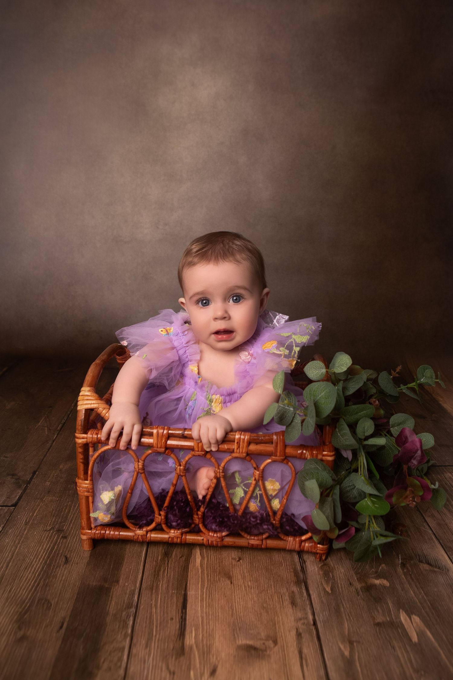 Baby girl in purple tutu sits in wicker basket with greenery on wooden floor against brown backdrop.