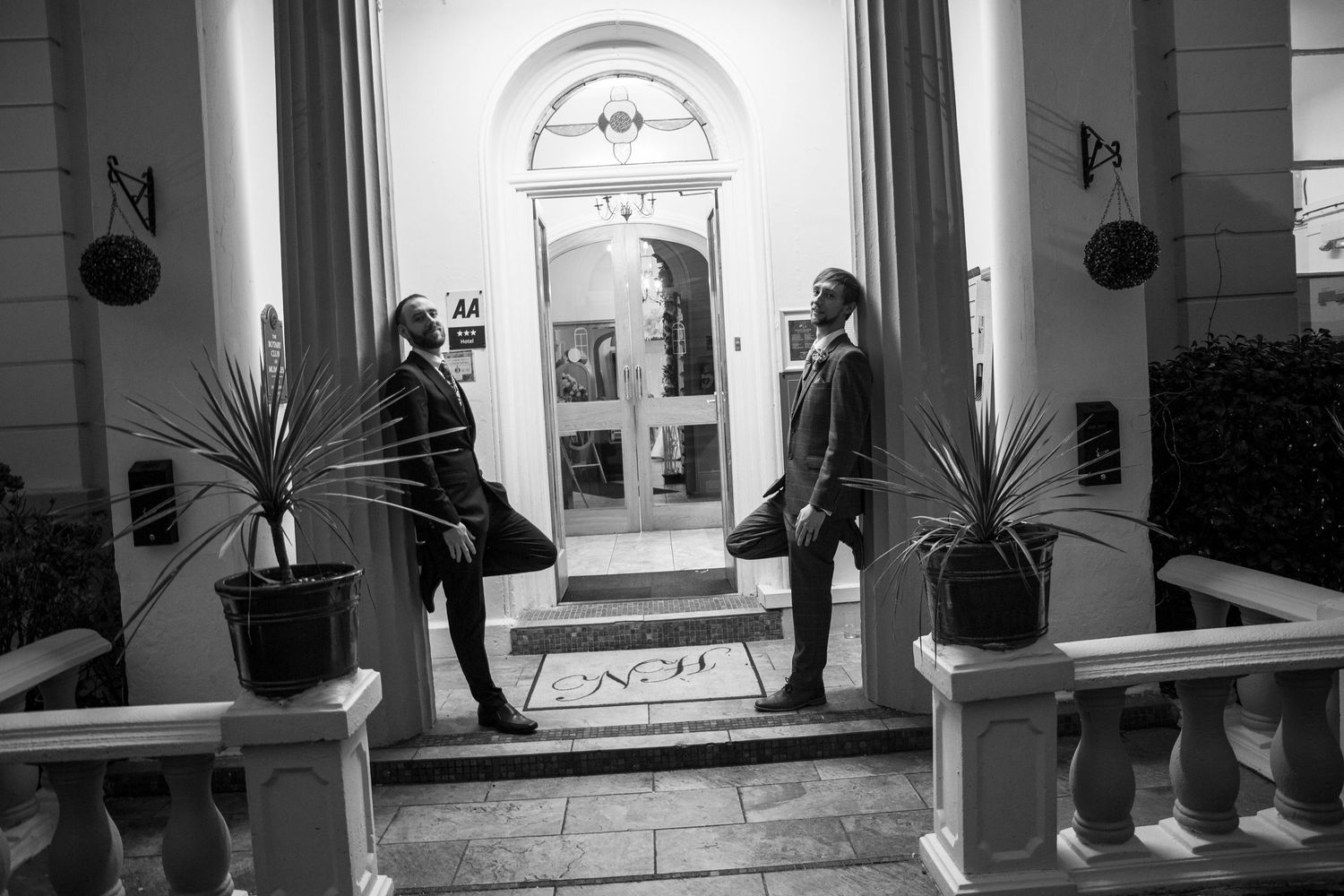 Two men in suits stand at a grand entrance with potted plants in a black and white photo.