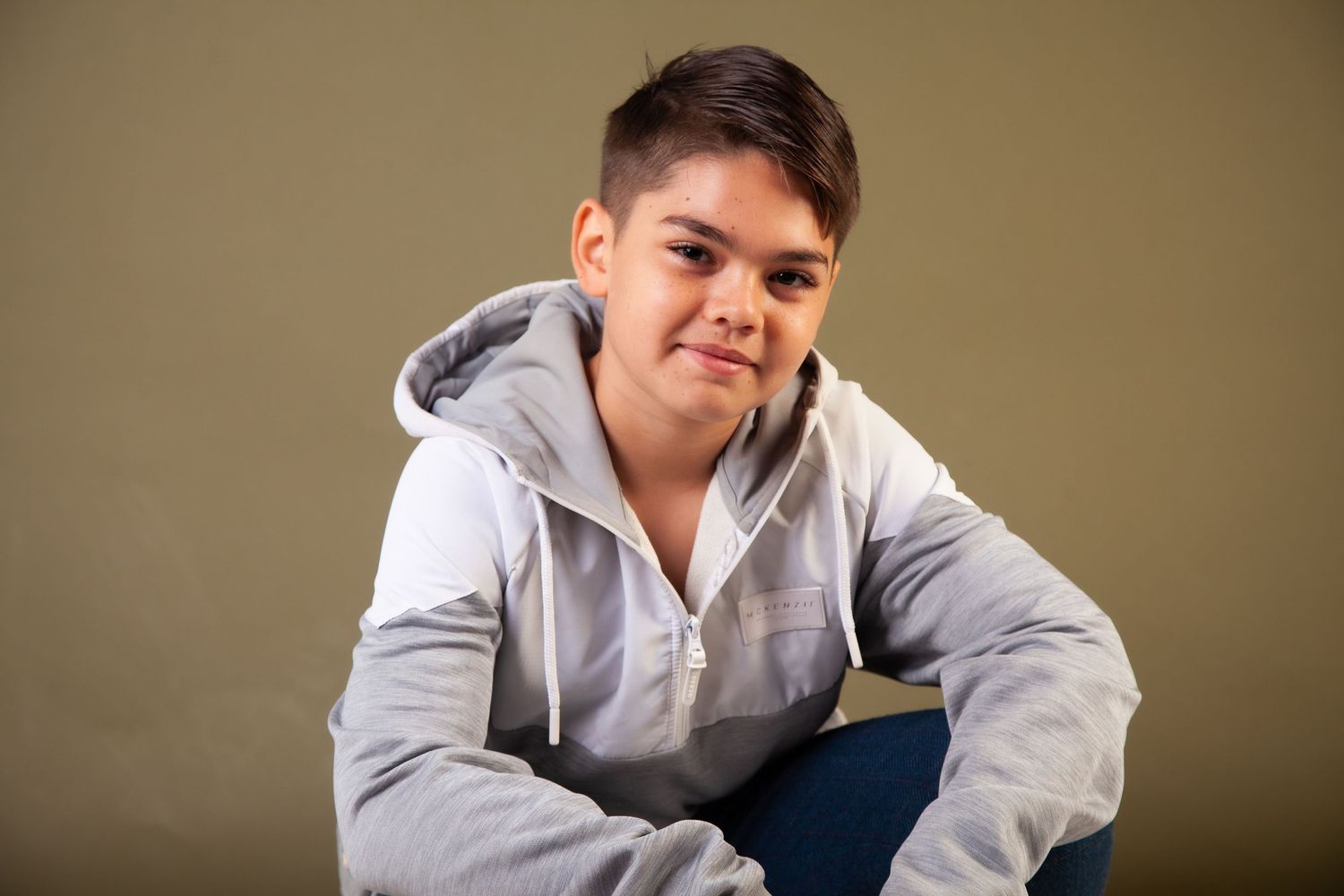 A teenage boy in a gray hoodie sits casually against a tan background with a relaxed expression.