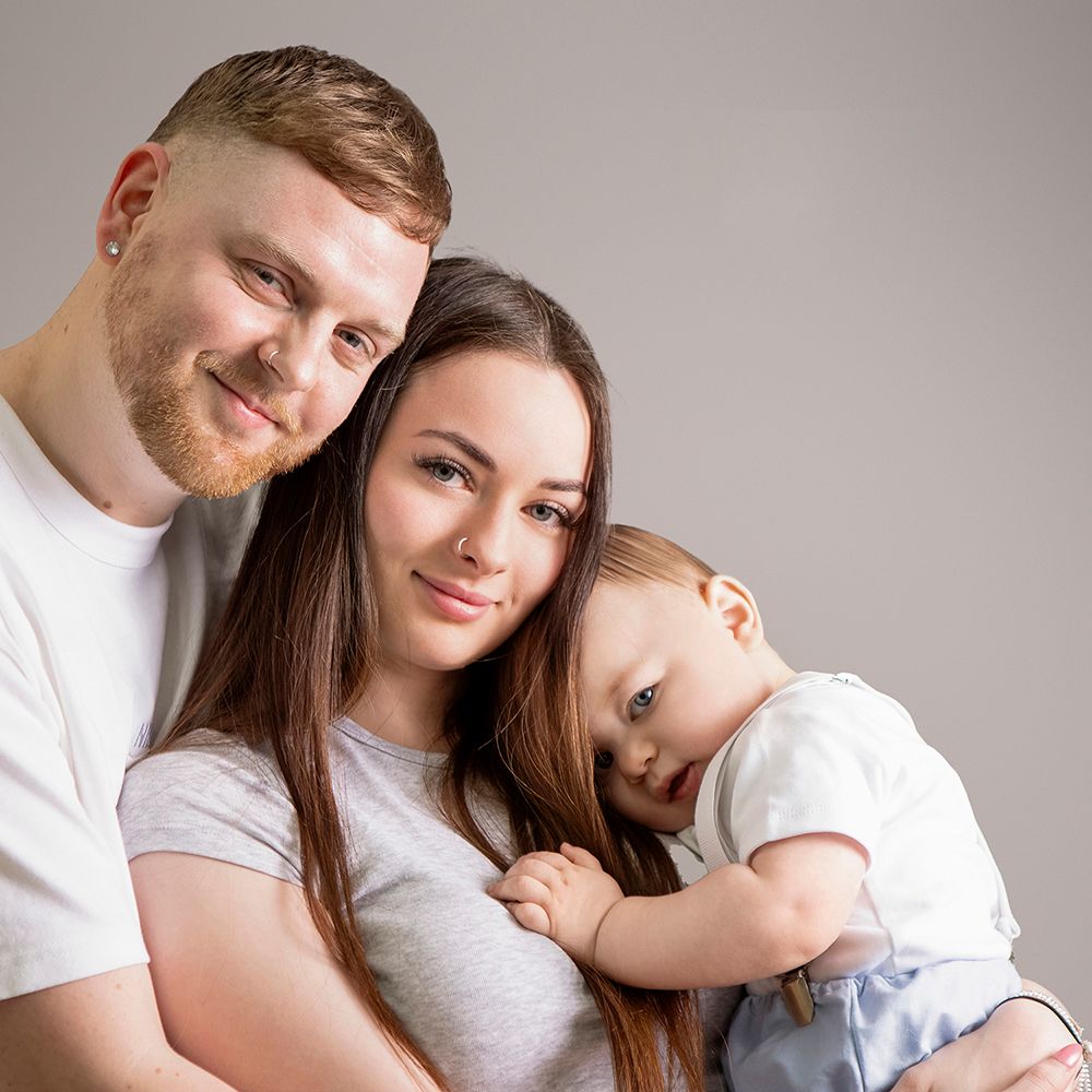 A young family poses for a studio portrait, with parents smiling and holding their sleepy toddler in a white outfit.