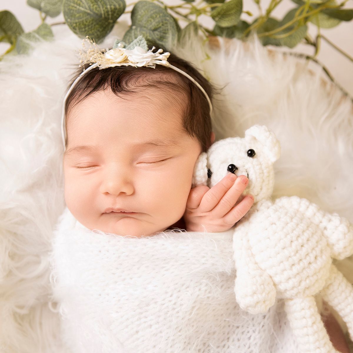 Sleeping newborn baby wrapped in white, holding a crochet teddy bear, wearing a floral headband on a fluffy white blanket.