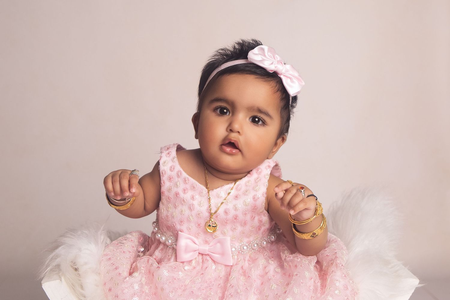 Adorable baby girl in pink polka dot dress with gold jewelry and pink bow headband posed for portrait photo.