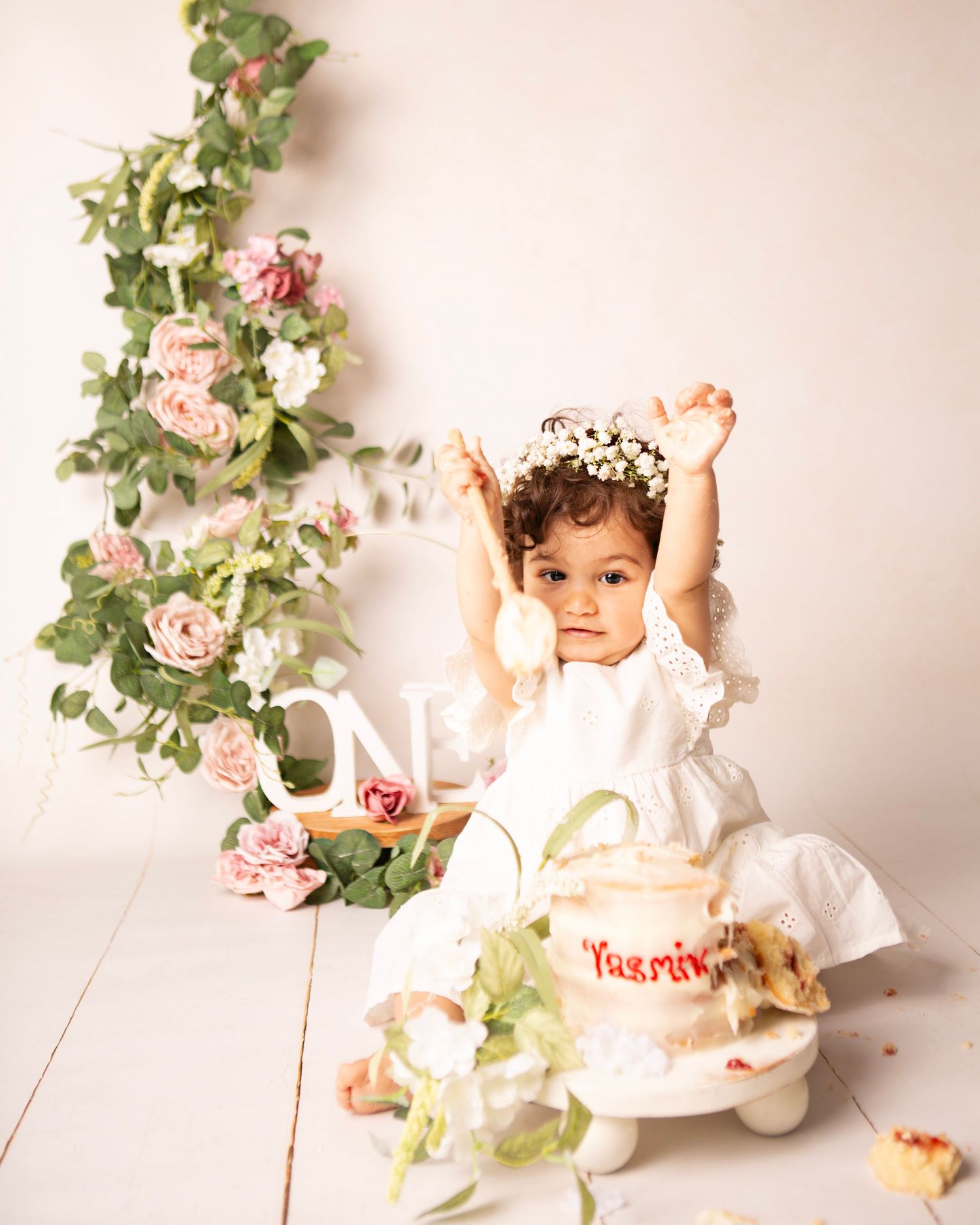 Baby girl in white dress with floral crown celebrates first birthday smashing cake surrounded by pink roses.