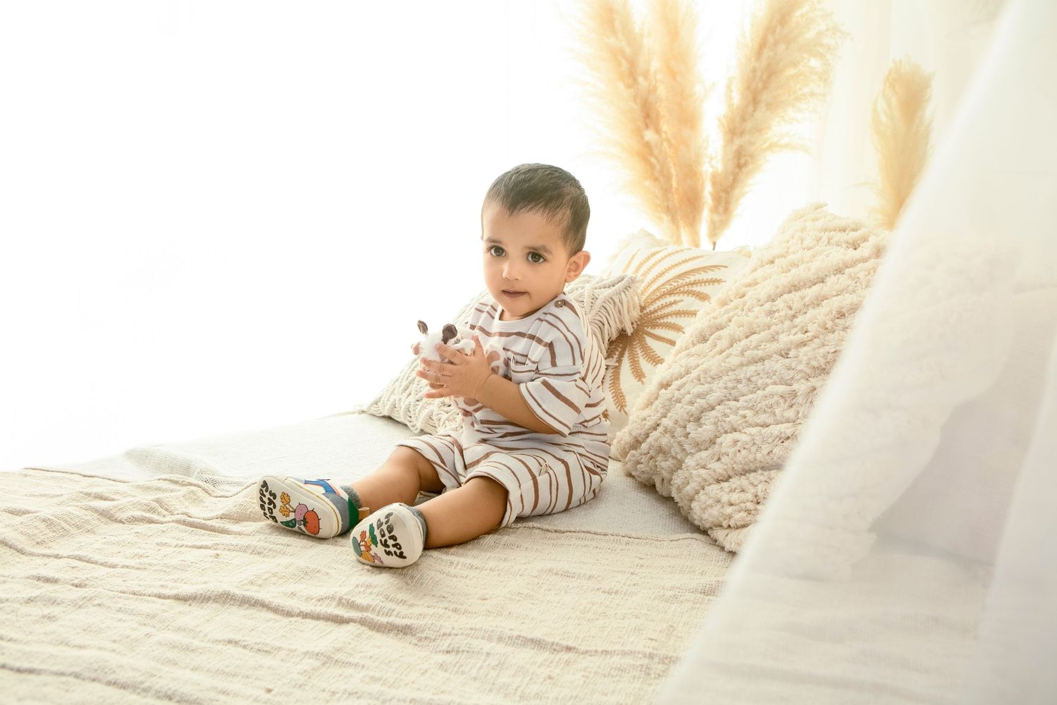 Adorable baby boy sitting on a white knit blanket in a boho nursery, wearing striped outfit and colorful shoes.