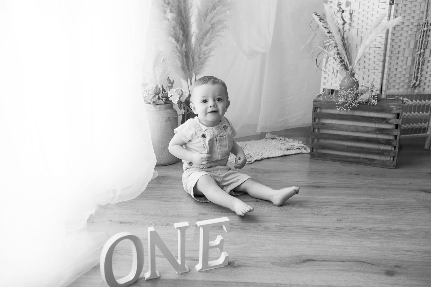 Black and white photo of a baby sitting on floor beside ONE letters sign, celebrating first birthday.