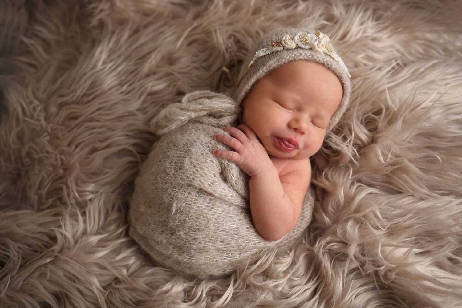 Peaceful newborn baby swaddled in gray wrap wearing knit bonnet, sleeping on fluffy beige fur blanket.