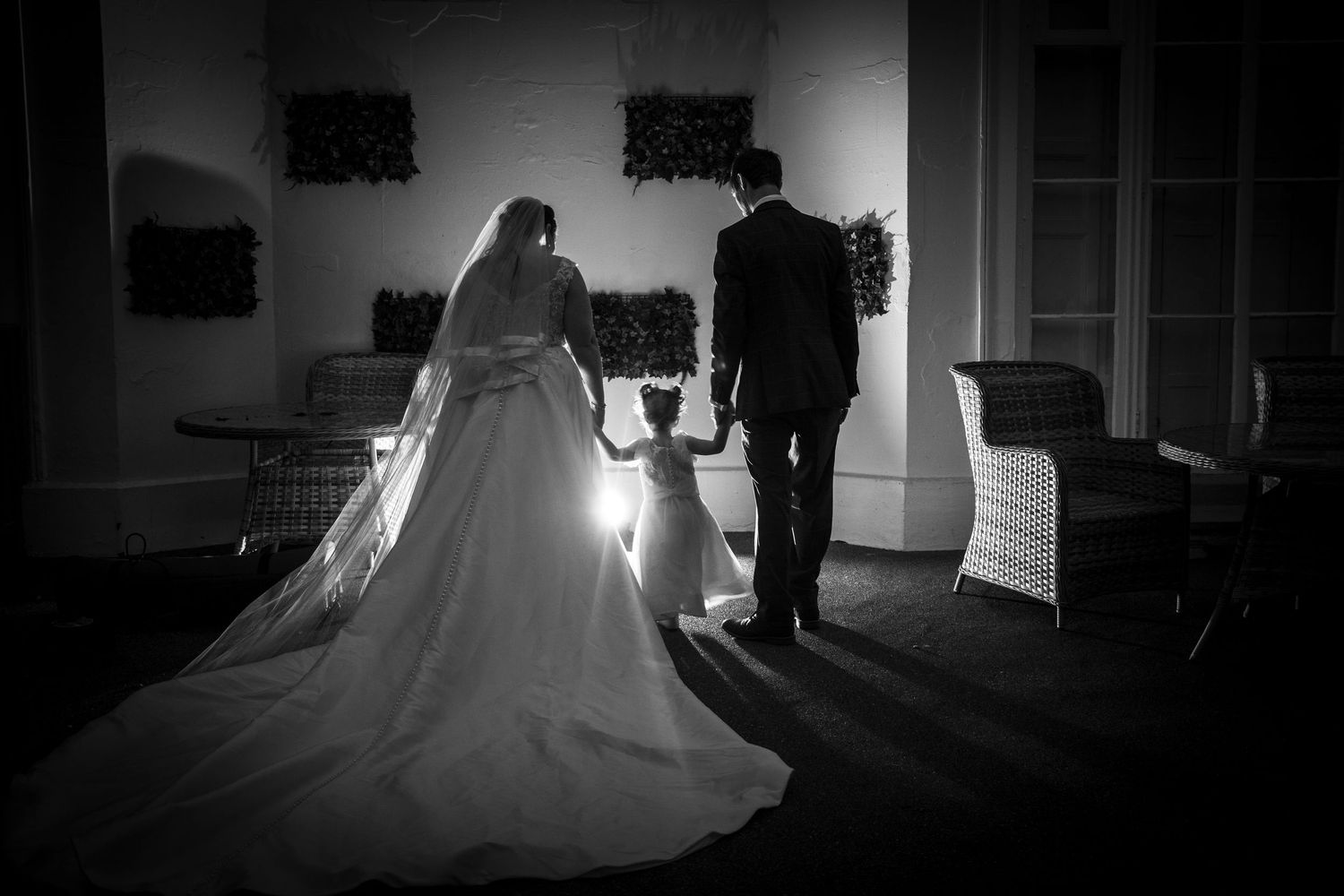 Black and white wedding photo of bride and groom holding hands with flower girl, elegant gown with long veil.