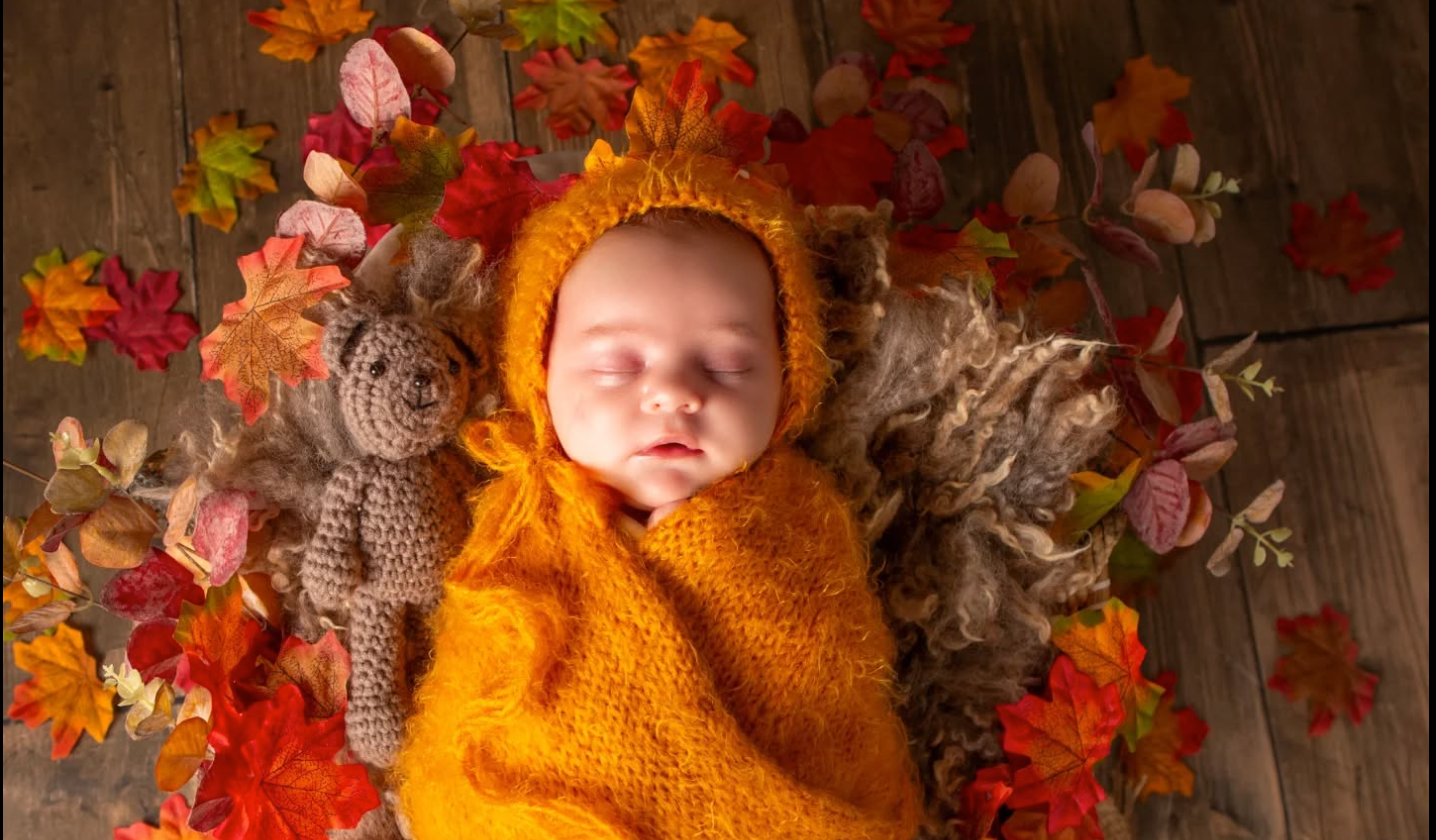 Newborn baby wrapped in orange knit swaddle with bear bonnet, surrounded by colorful autumn leaves and a crochet teddy bear.