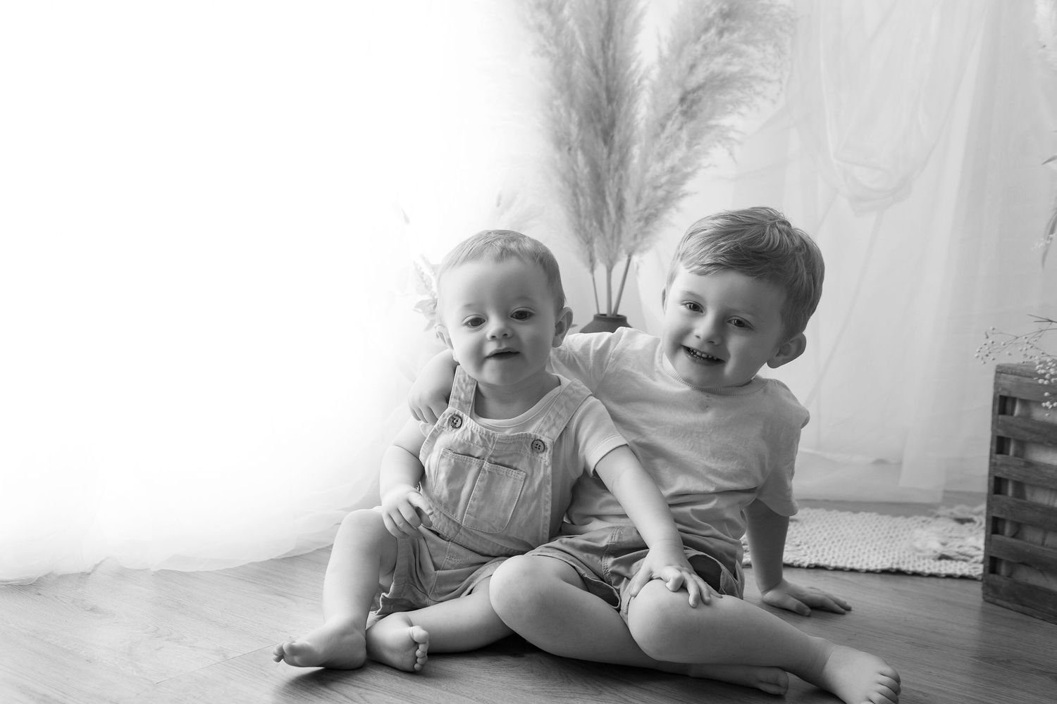Two happy toddlers sitting together on a wooden floor in a black and white portrait photo.