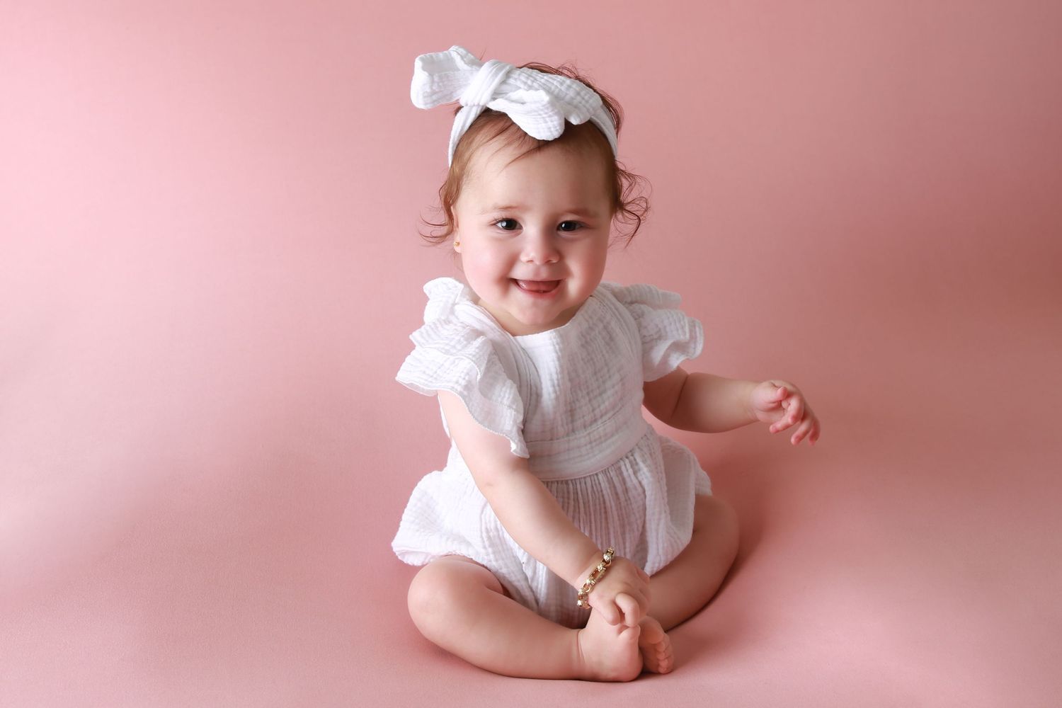 Smiling baby girl in white dress and bow headband sitting on pink background.