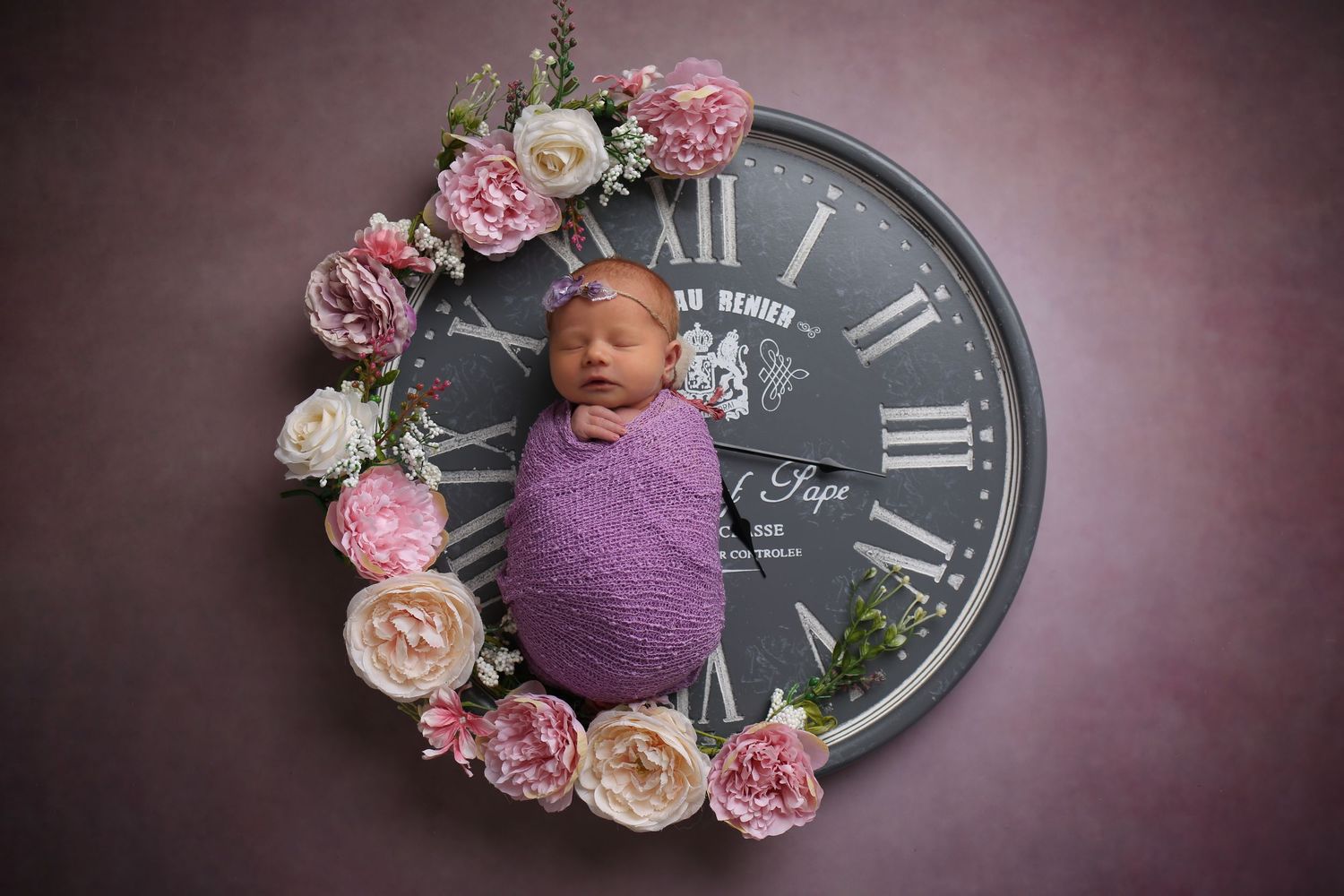 Newborn baby swaddled in purple wrap sleeping on a floral clock prop with pink and white roses.