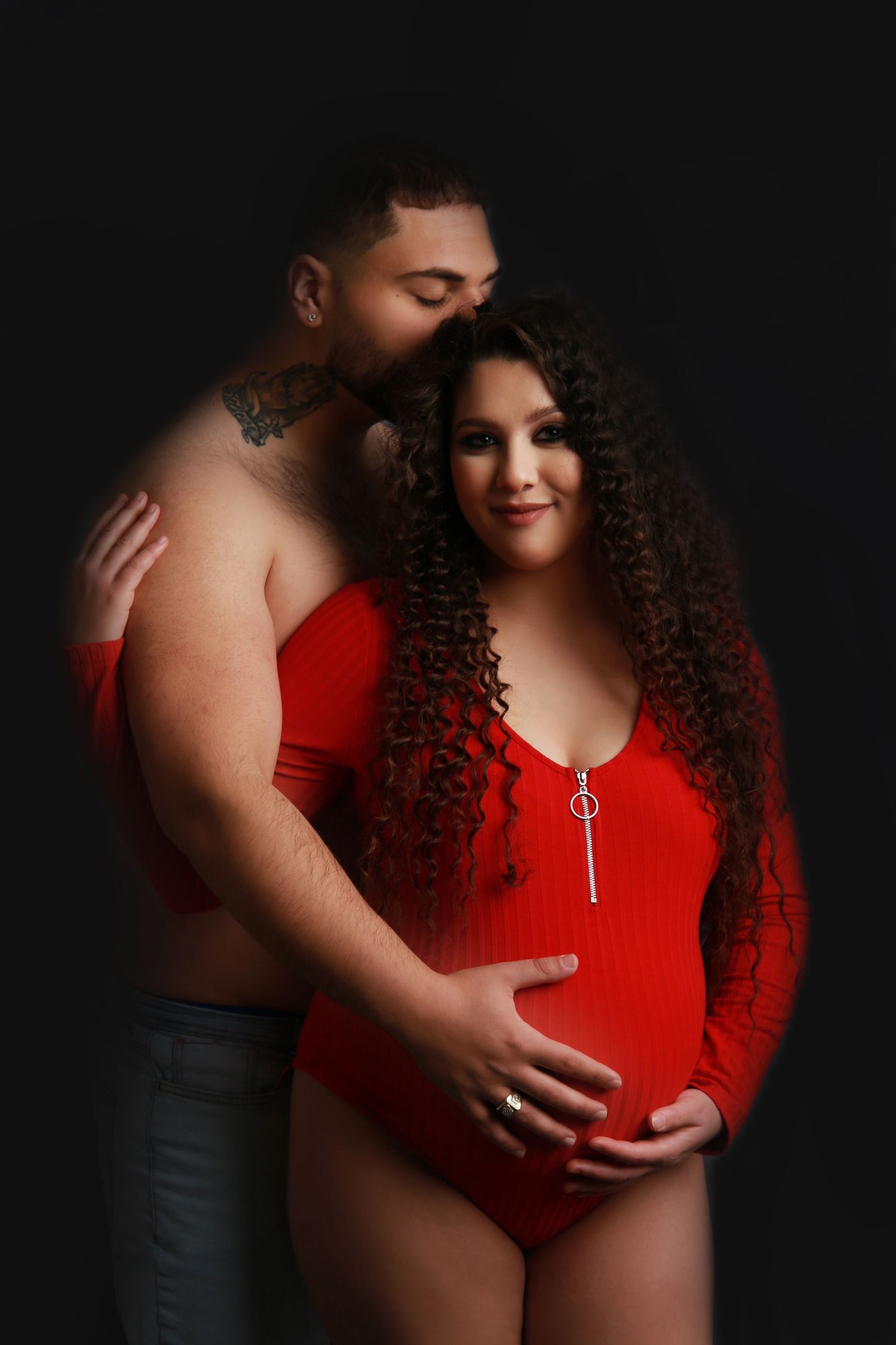 Couple maternity photo with tattooed shirtless man and pregnant woman in red bodysuit.