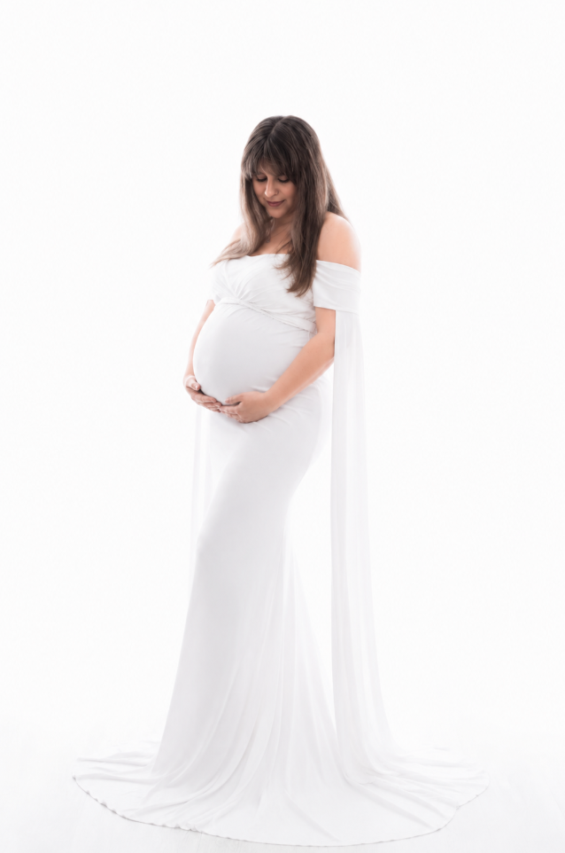 Elegant pregnant woman in white off-shoulder gown with flowing train gazing down at her belly.