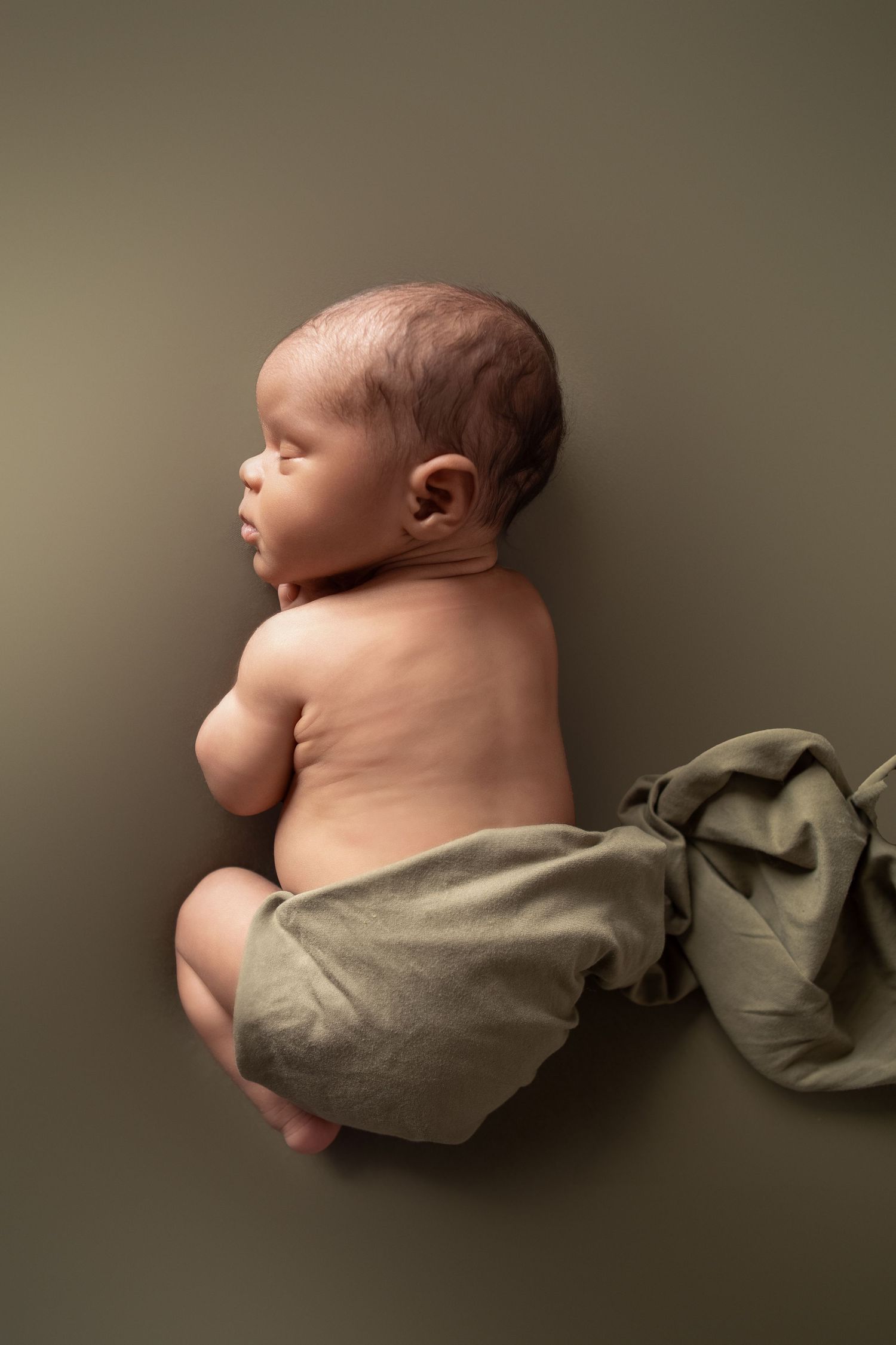 Newborn baby lying on side wrapped in olive green fabric against matching background.