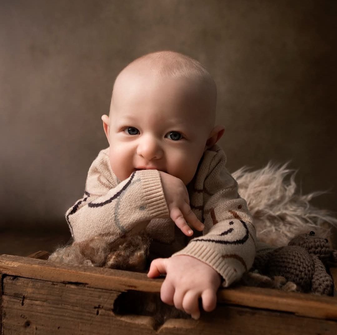 Adorable baby in a beige knit sweater lying in a rustic wooden box, smiling with hand near mouth.