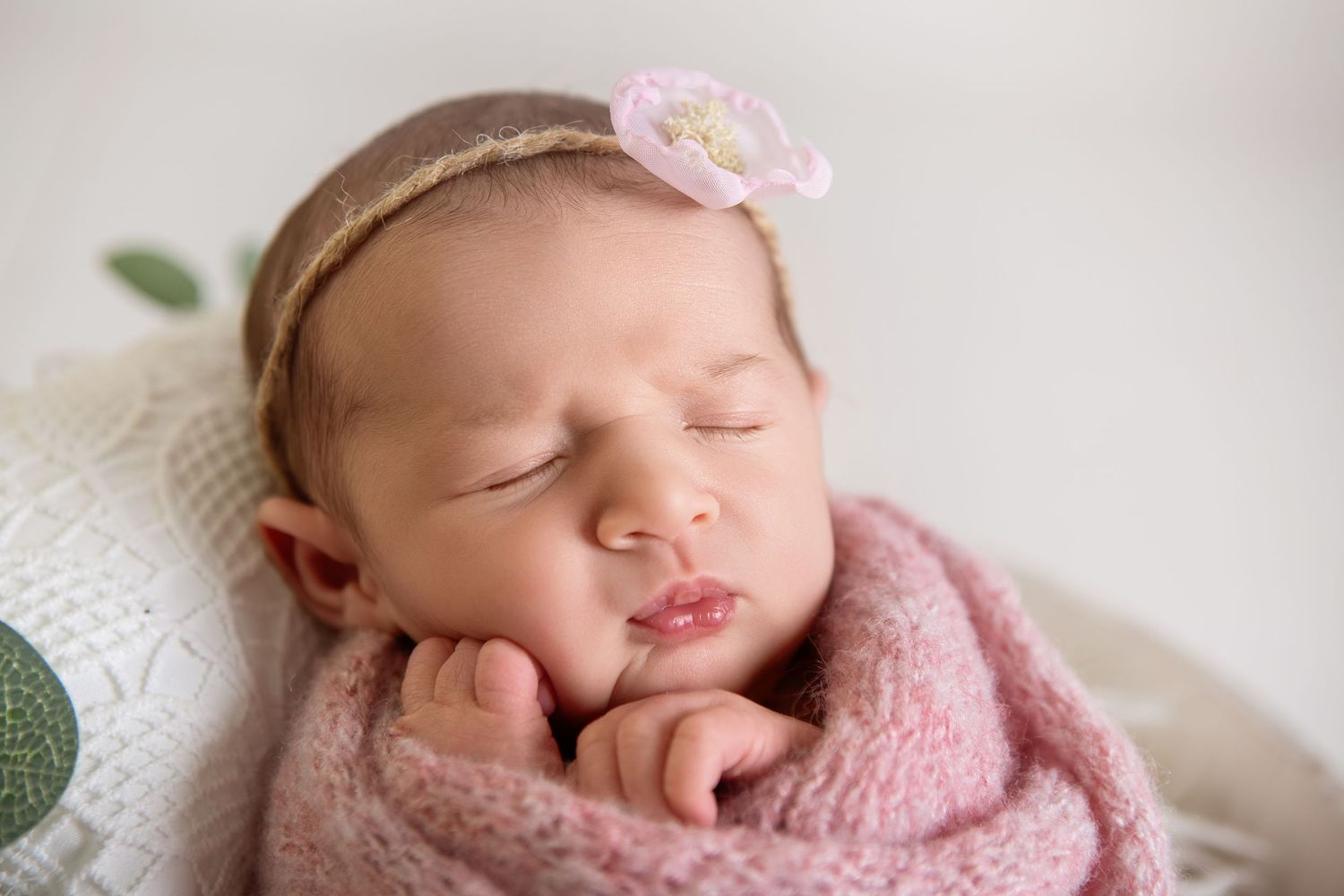 Peaceful newborn baby girl wrapped in pink knit blanket wearing a floral headband sleeping with hands near face.