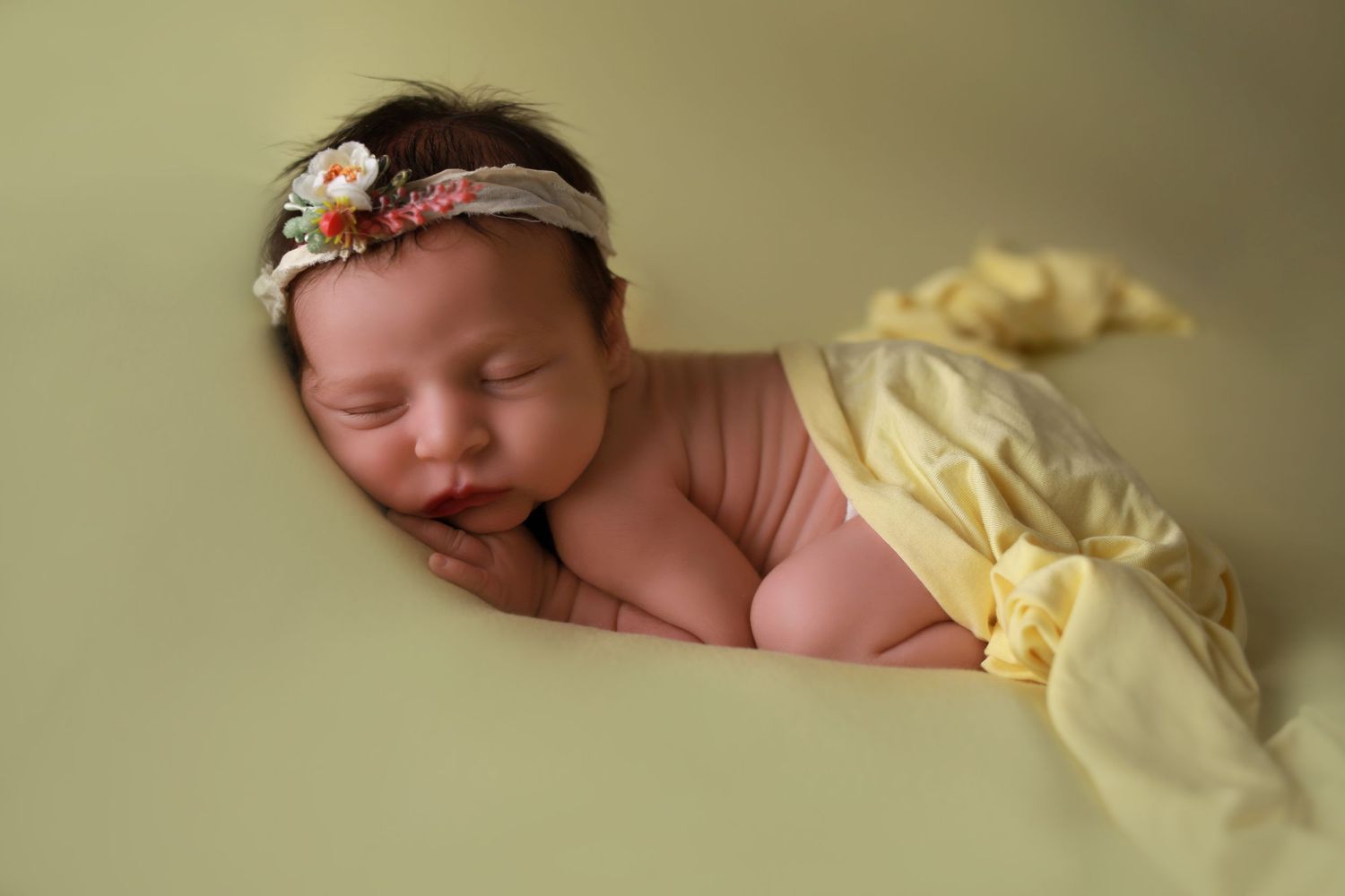 Peaceful newborn baby girl sleeping on green backdrop wearing floral headband wrapped in yellow fabric.