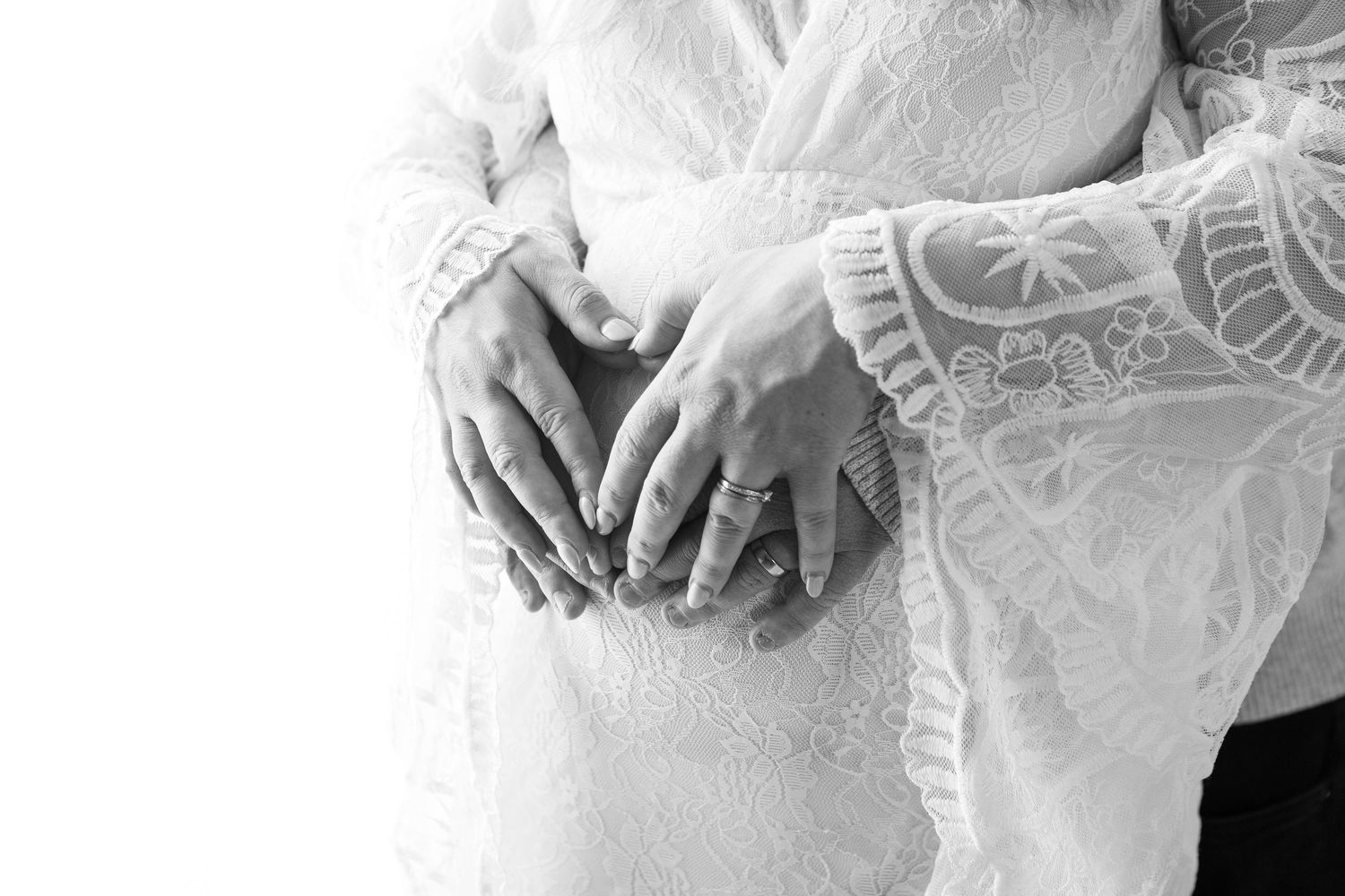 Black and white close-up of couple's hands forming heart shape on pregnant belly in lace dress.