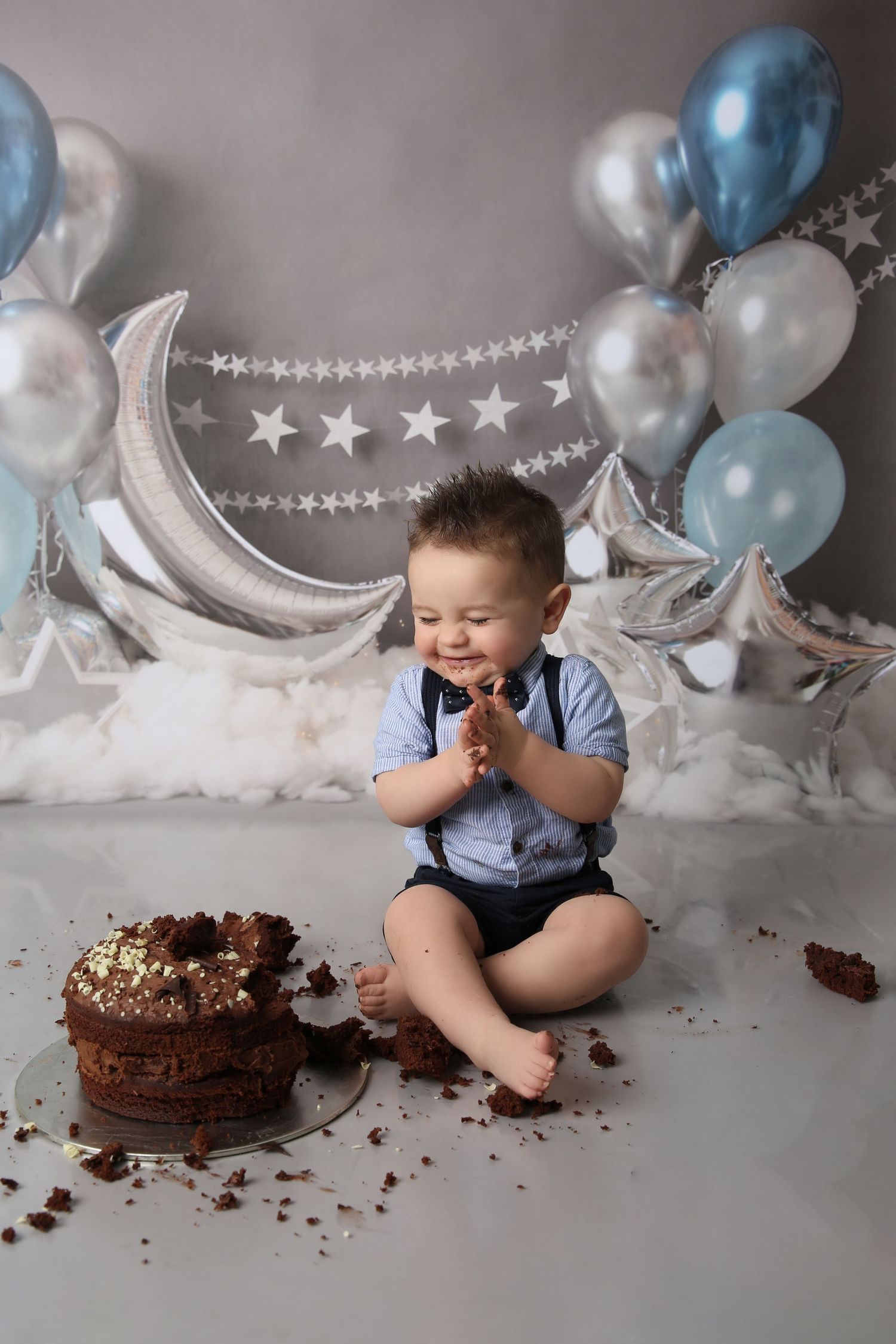 Smiling baby boy in blue outfit enjoys first birthday cake smash with balloons and moon decorations.
