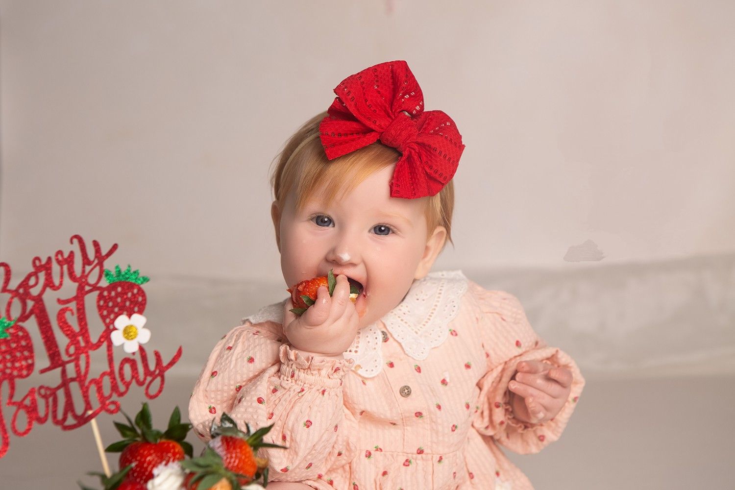 Baby girl in pink dress with red bow eating strawberry cake at her Berry 1st Birthday party celebration.