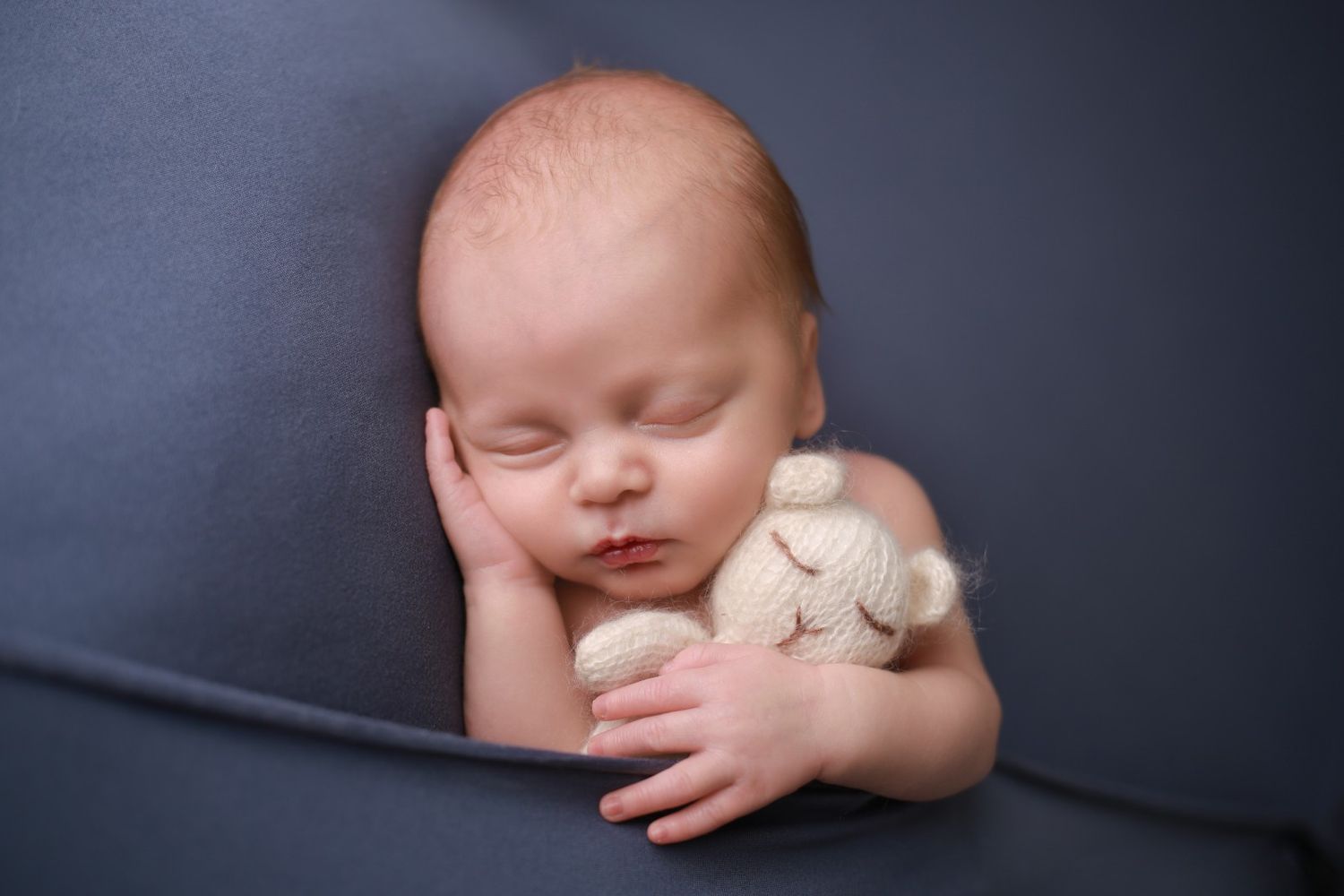 Newborn baby sleeping peacefully on gray fabric, cuddling a small knit stuffed animal toy.