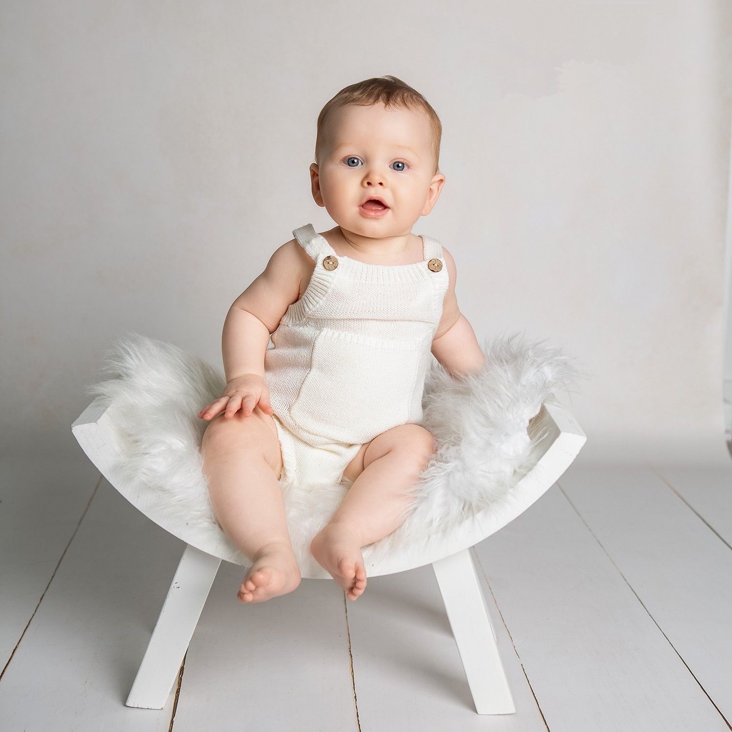 A cute baby in a white romper sits on a small white wooden chair with fluffy white fur trim in a studio setting.
