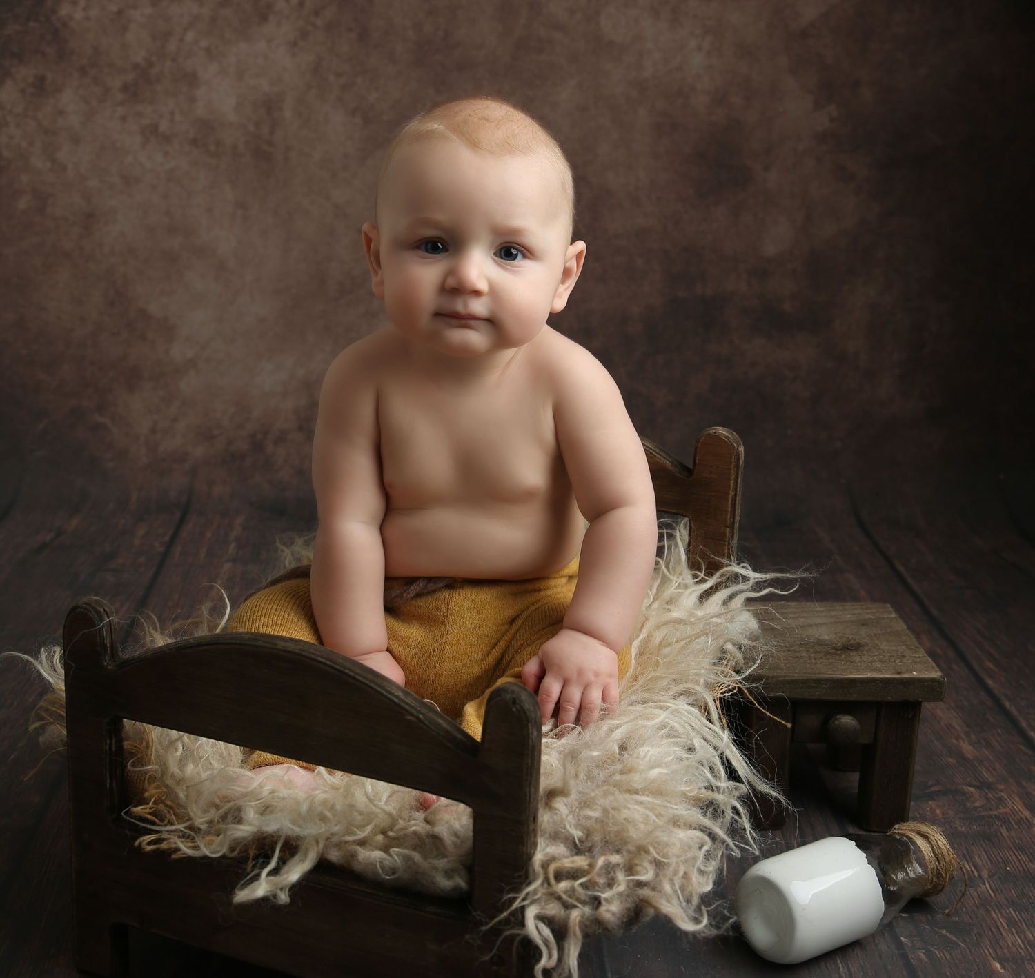 Adorable baby sitting in a rustic wooden bed with fluffy wool, wearing mustard shorts, in a professional studio portrait.