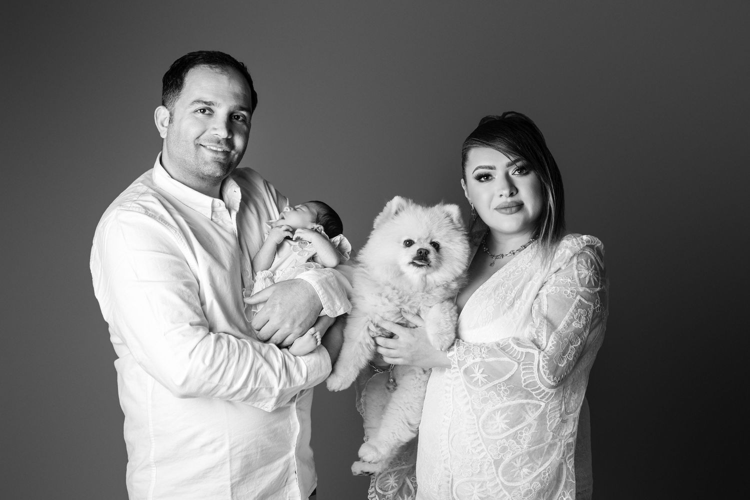 Black and white family portrait of couple holding newborn baby and fluffy white Pomeranian dog in studio.