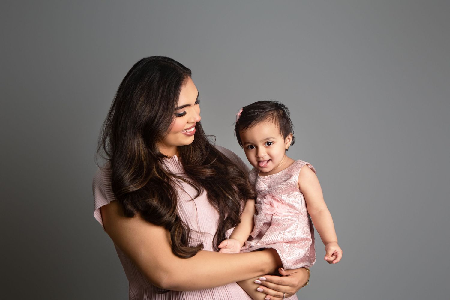 A young woman in pink dress holds a smiling baby girl, both wearing pink, against a gray studio background.