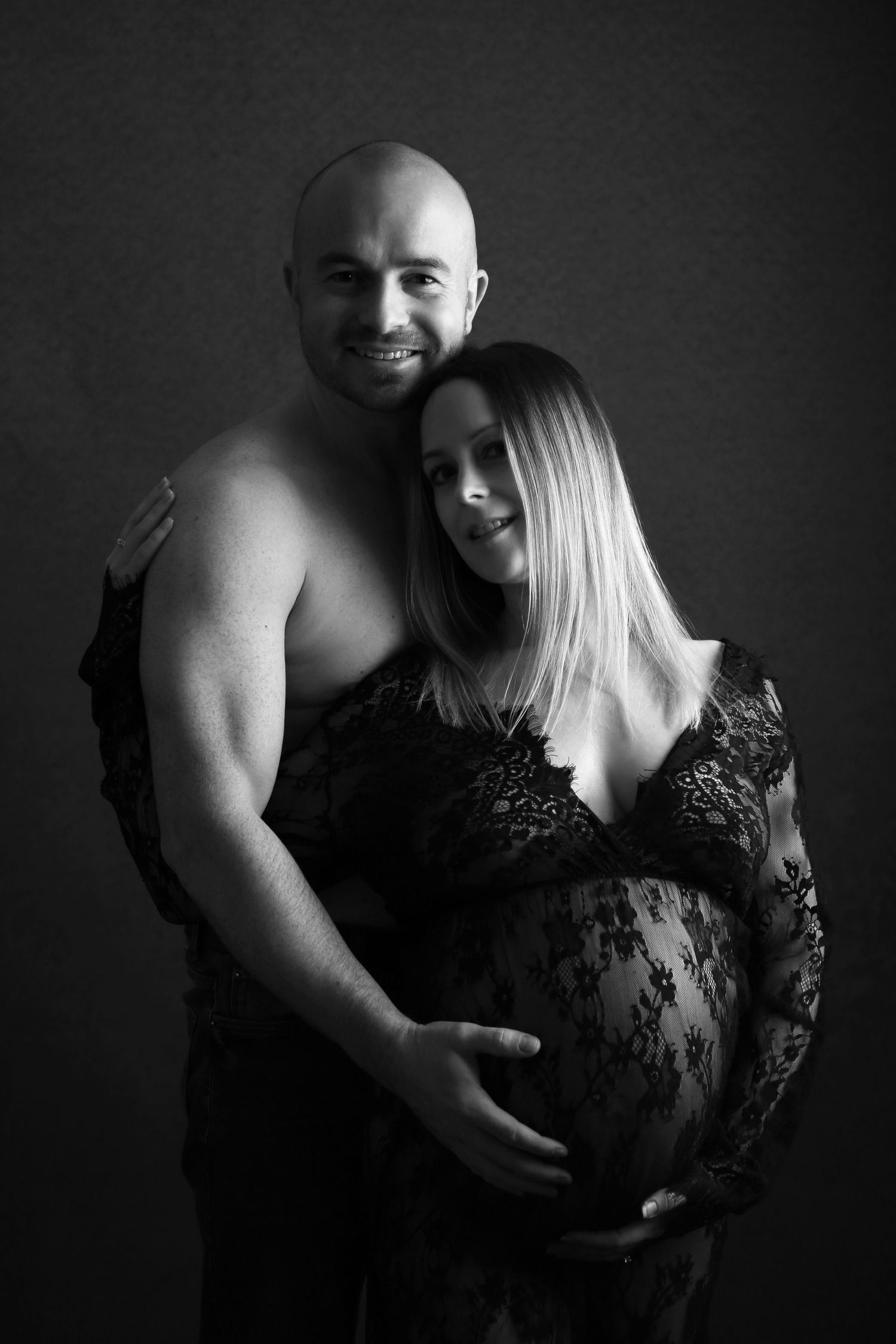 Black and white maternity photo of a shirtless man embracing a pregnant woman in lace dress, both smiling softly.