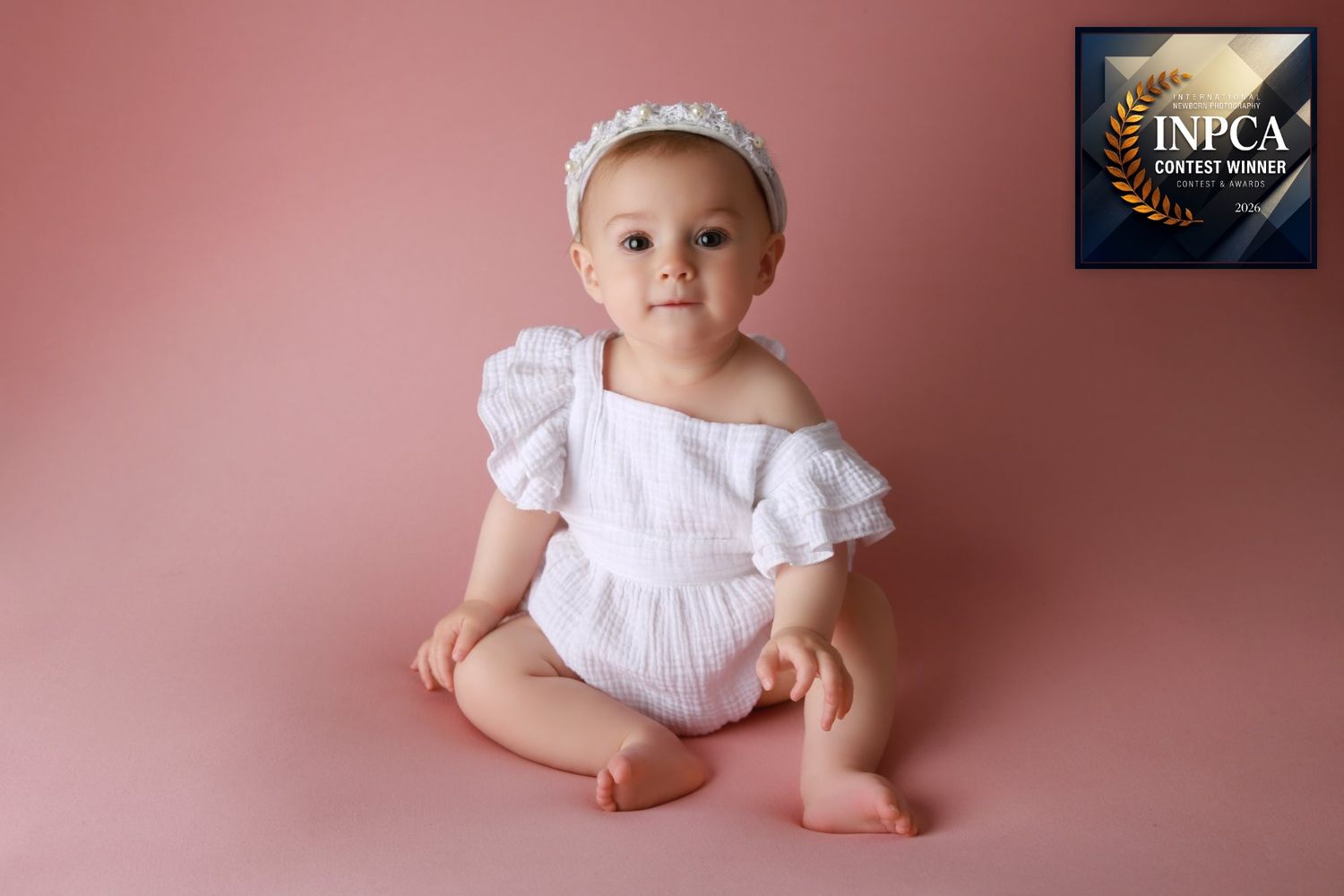 An award winning portrait of a confident 6 month old sitting independently against a soft pink backdrop.