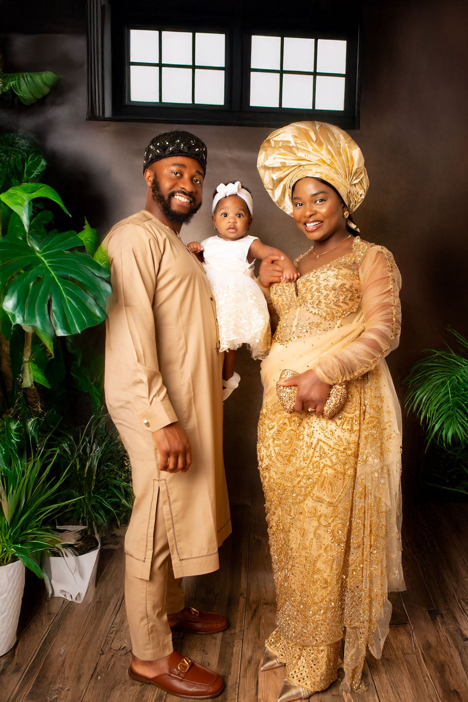 Nigerian family in traditional gold attire posing with baby in white dress for a formal portrait photo.