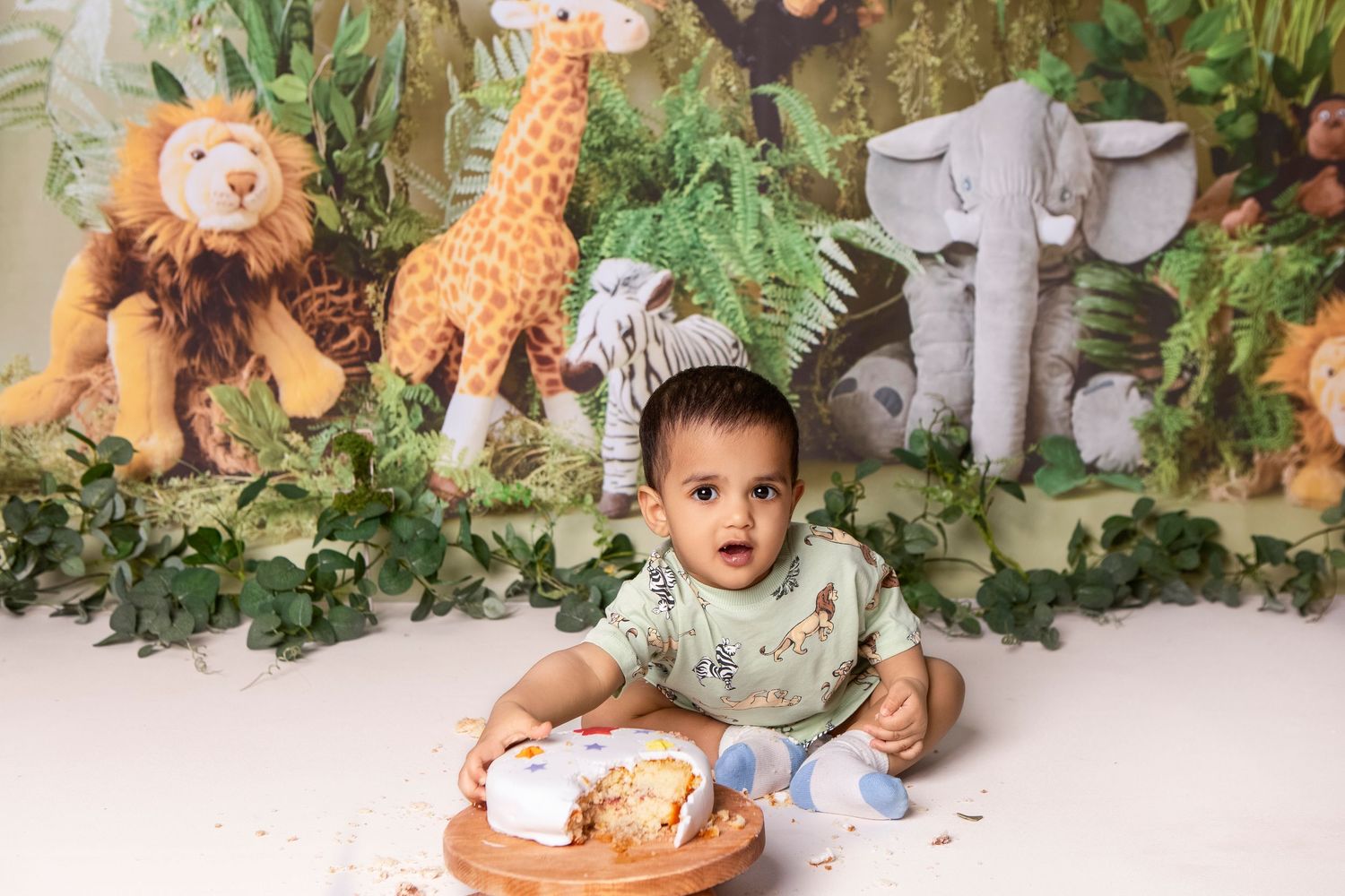 Baby's first birthday cake smash with jungle safari theme, stuffed animal toys, and lush green backdrop.