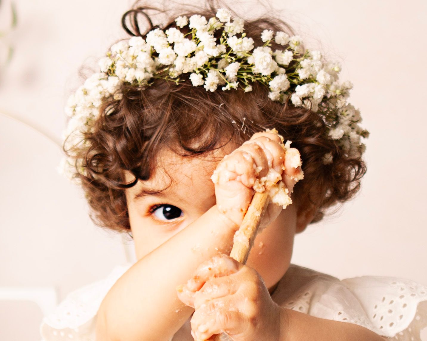 Toddler with curly hair wearing a white flower crown holds a piece of cake, with frosting on hands.