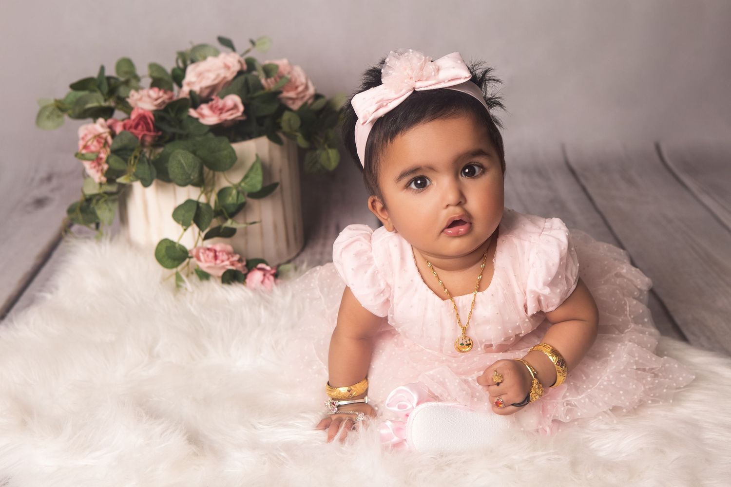 Adorable baby girl in pink ruffle dress and gold jewelry poses on fluffy white rug with pink roses backdrop.