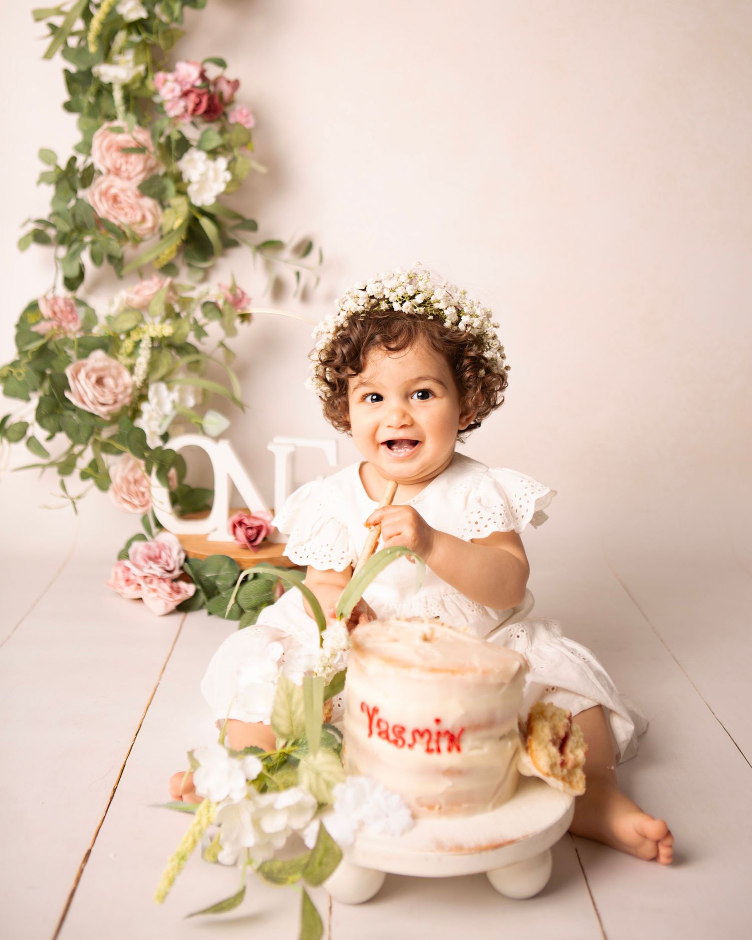 Smiling baby girl in white dress and flower crown sits beside a personalized first birthday cake with pink floral backdrop.