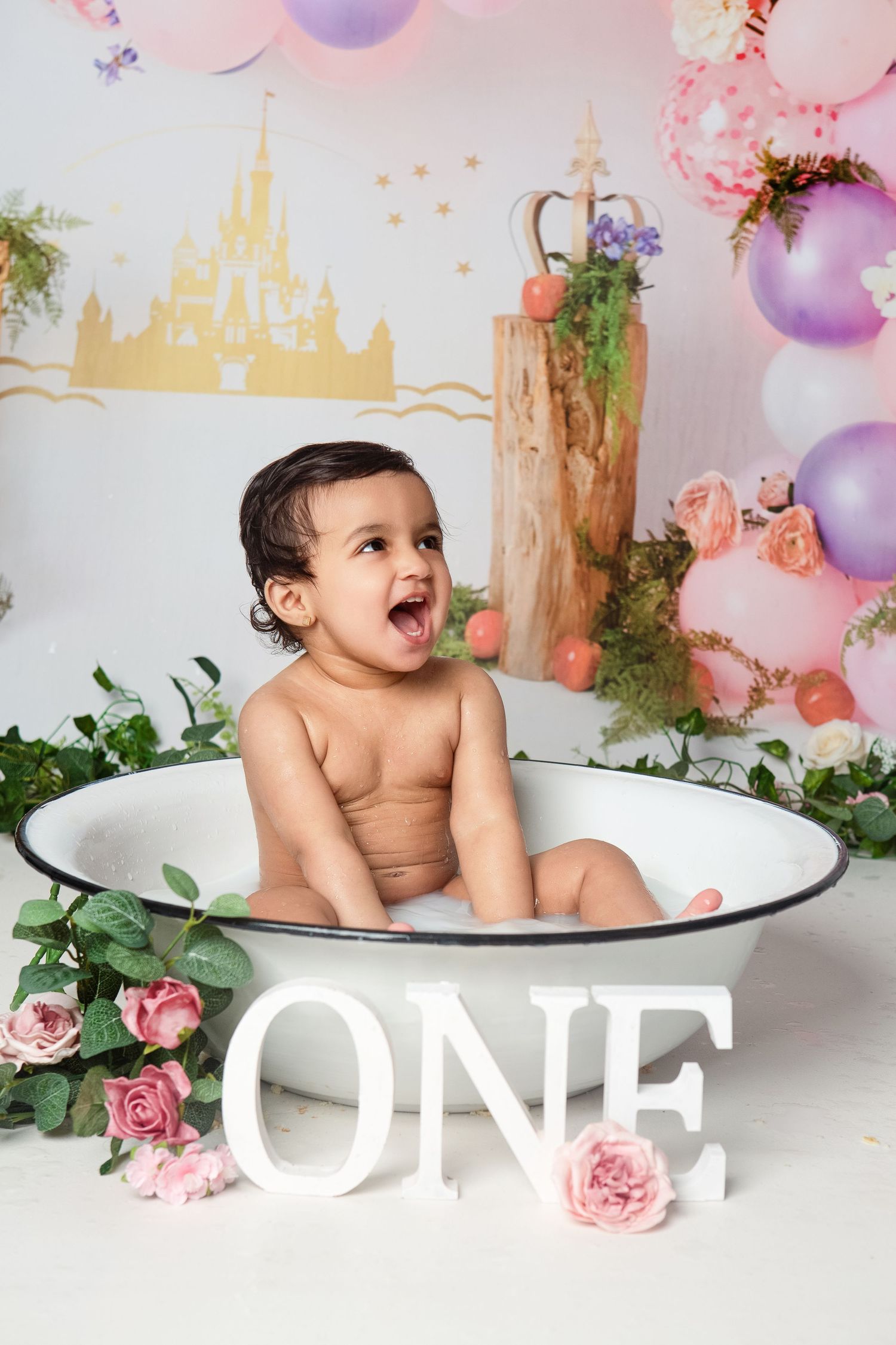 A happy baby sits in a vintage tub surrounded by flowers and balloons for a Disney-themed first birthday cake smash.
