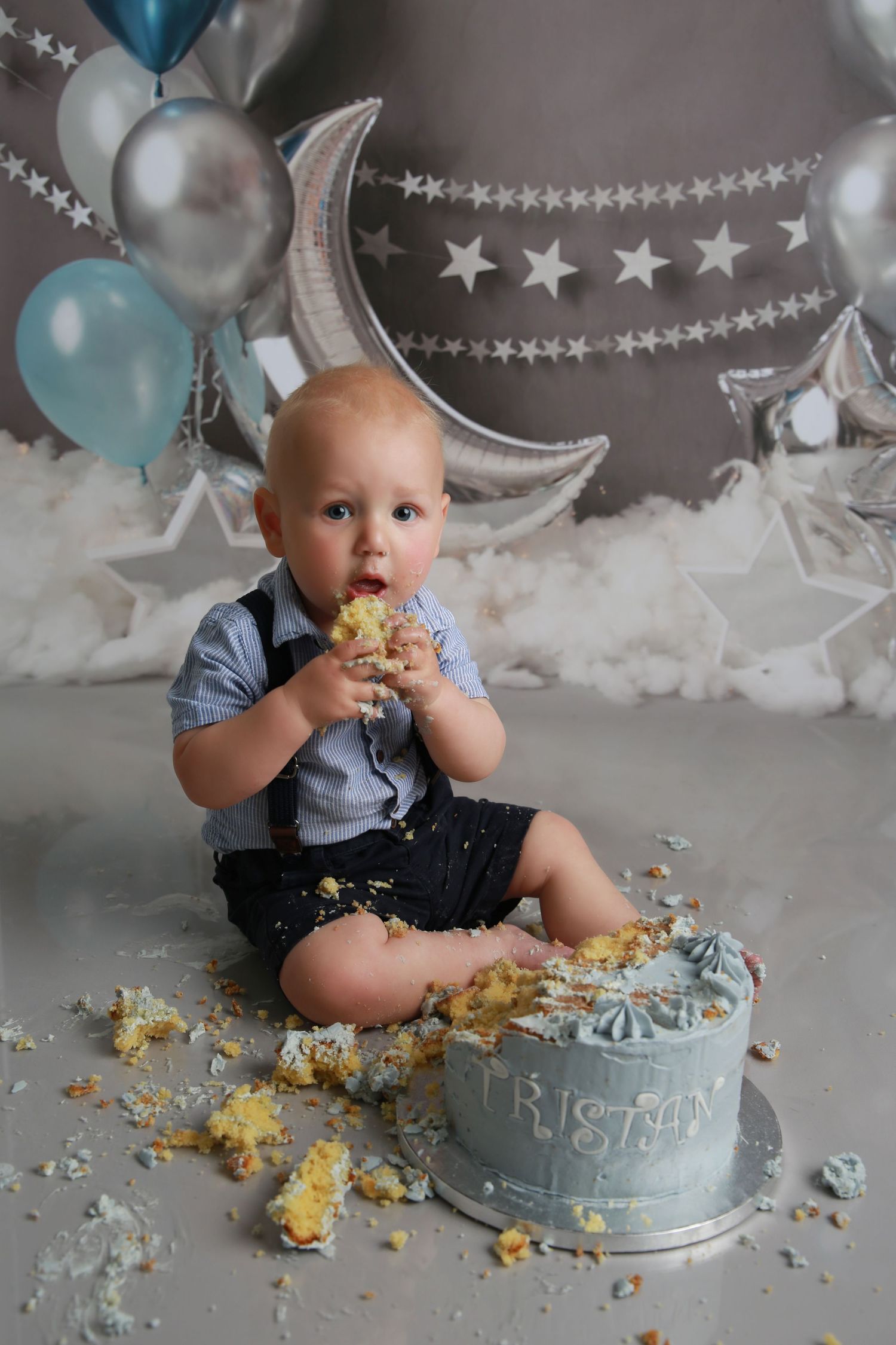 Baby boy smashing his first birthday cake surrounded by silver and blue balloons, stars, and moon decorations.