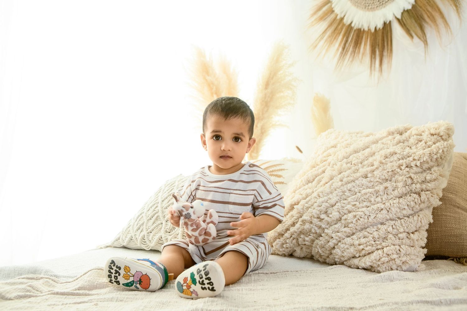 Cute toddler boy sitting on a bed with cream pillows wearing a striped outfit and colorful shoes.