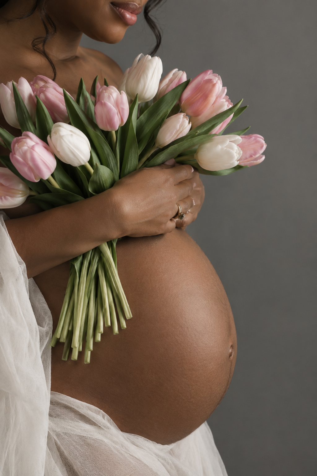 Pregnant woman holding pink and white tulip bouquet against her bare belly, wearing white sheer fabric.