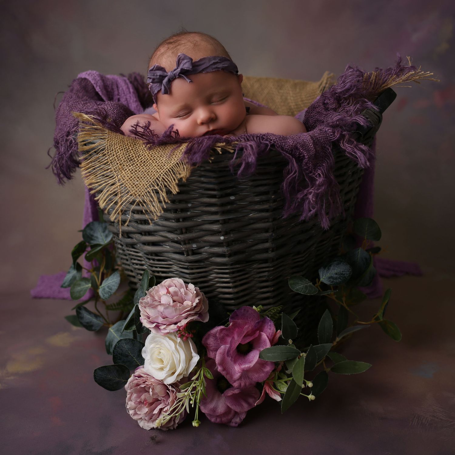 Sleeping newborn baby in purple wrap and floral headband resting in a wicker basket with purple flowers.
