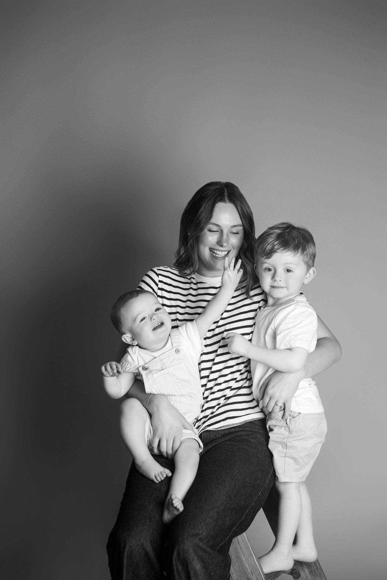 Black and white photo of a smiling mother in striped shirt holding two young boys in a studio portrait.