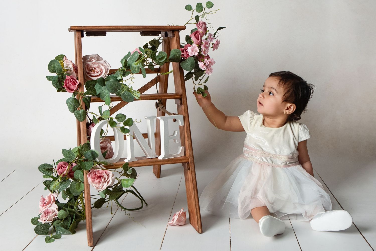 Toddler girl in white dress reaching for pink roses on a wooden ladder decorated with ONE sign for first birthday.
