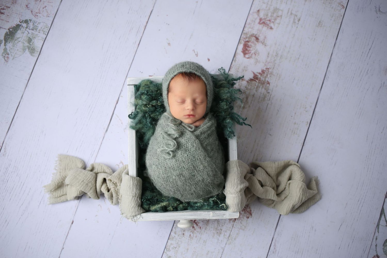 Newborn baby swaddled in green wrap sleeping in a white wooden box with matching green bonnet and fabric.