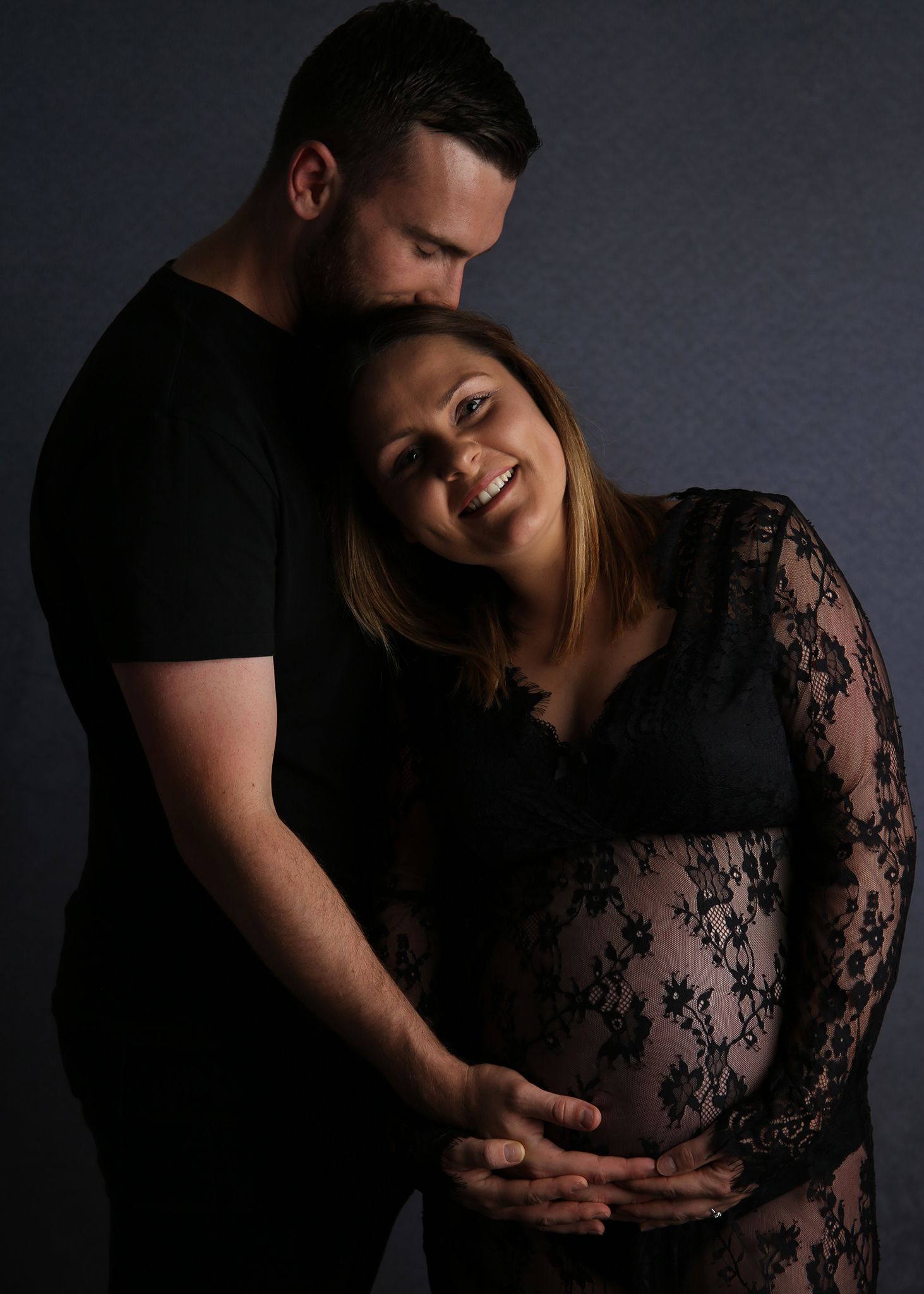 Couple in black outfits posing for a maternity photo, man embracing pregnant woman from behind in studio.
