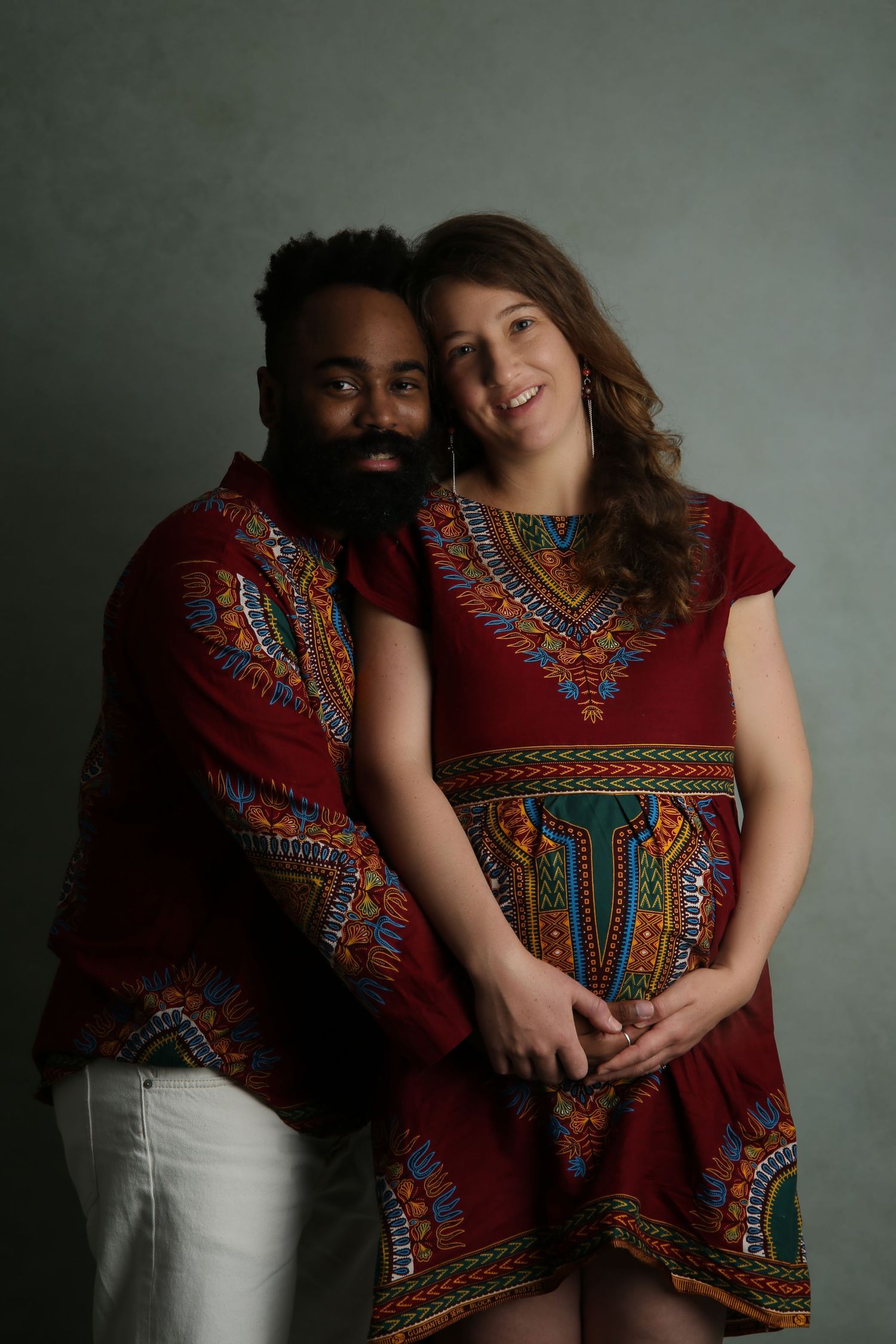 Couple wearing matching red dashiki outfits posing together, smiling warmly in a studio portrait setting.